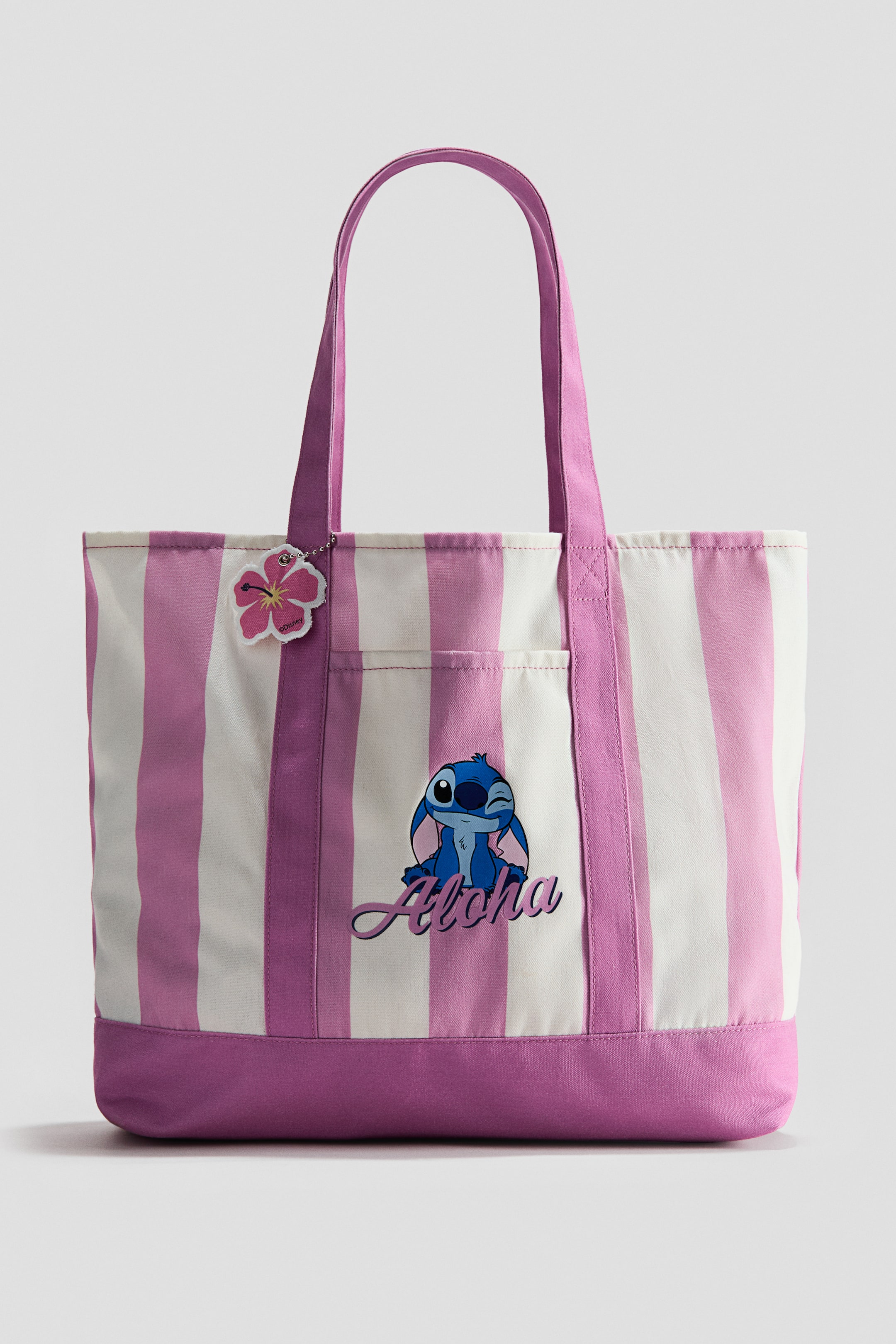 View larger image: A tote bag featuring vertical light pink and white stripes, with a solid fuchsia bottom panel and matching handles. The front displays a blue cartoon character, Stitch, winking, with the word 'Aloha' in script below him, alongside an exterior slip pocket. A pink hibiscus flower charm hangs from one of the handles.