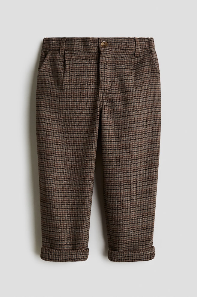 The front-facing brown and black houndstooth pattern pants are laid flat, featuring a button closure on the waistband, subtle pleats below the waistband, and rolled cuffs at the hem. - 1