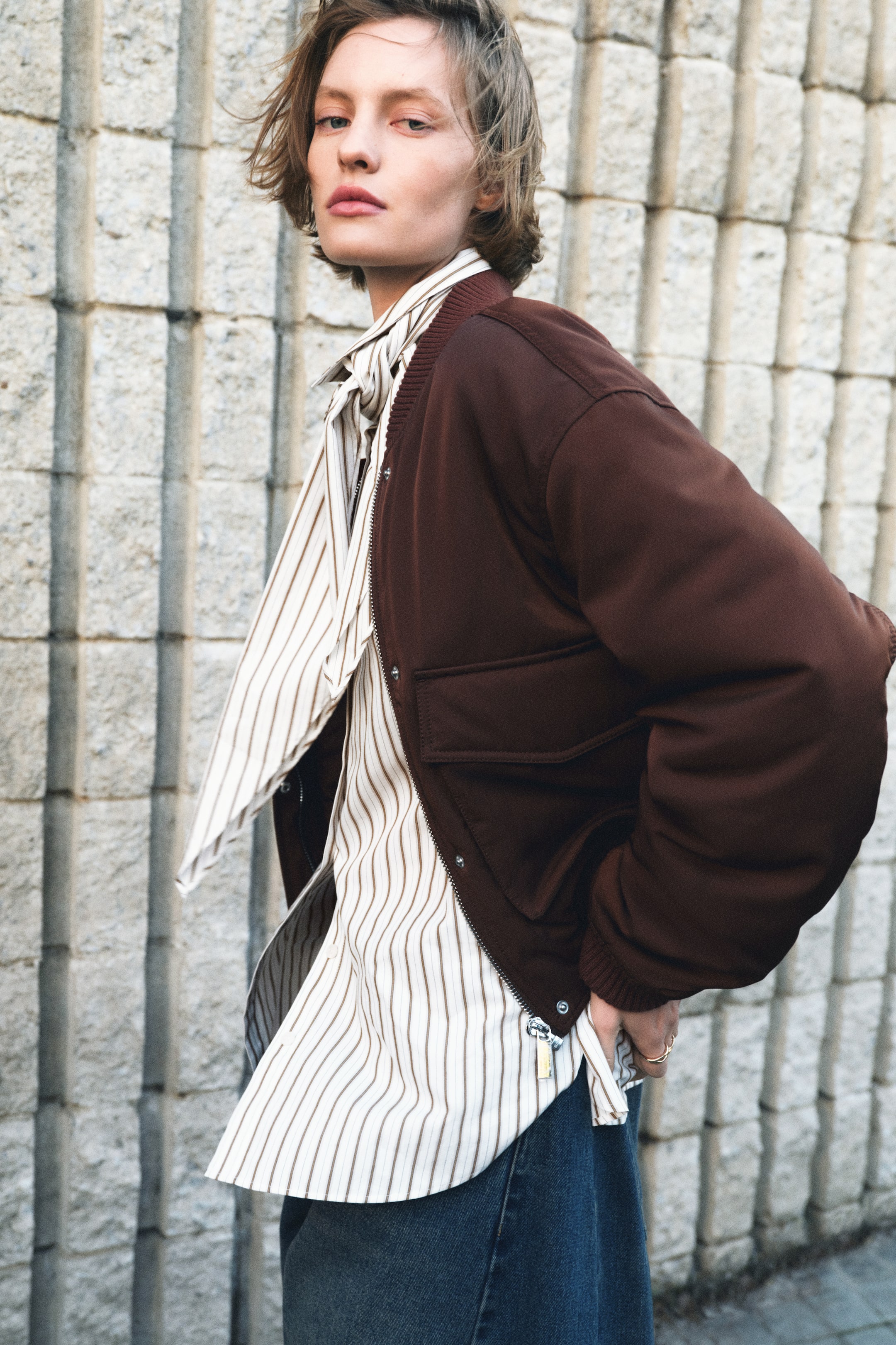 View larger image: The model is sporting a loose-fit, striped shirt crafted from woven fabric, featuring a collar, buttons running down the front, a yoke, dropped shoulders, long sleeves with buttoned cuffs, and a rounded hem. A matching striped scarf is elegantly tied around the model's neck, while the model's hand is casually tucked into the shirt's hem. The shirt is stylishly layered beneath an open brown bomber jacket.