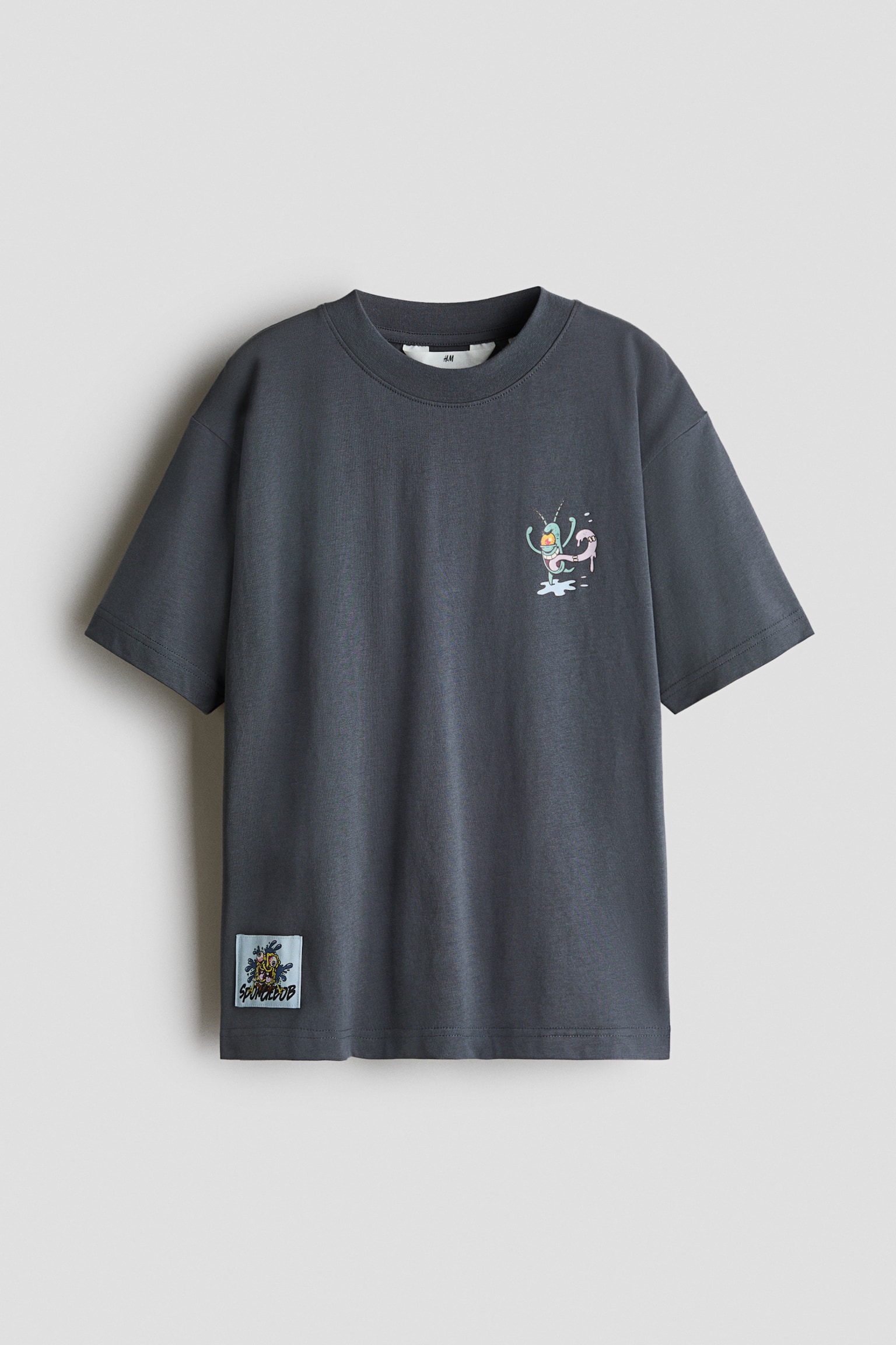 Un t-shirt en jersey de coton gris foncÃ© est prÃ©sentÃ© de face, avec un col rond bordÃ© de cÃ´tes et des Ã©paules tombantes. Il affiche un petit motif de dessin animÃ© imprimÃ© sur la poitrine ainsi quâun Ã©cusson rectangulaire bleu pÃ¢le prÃ¨s de lâourlet du bas.