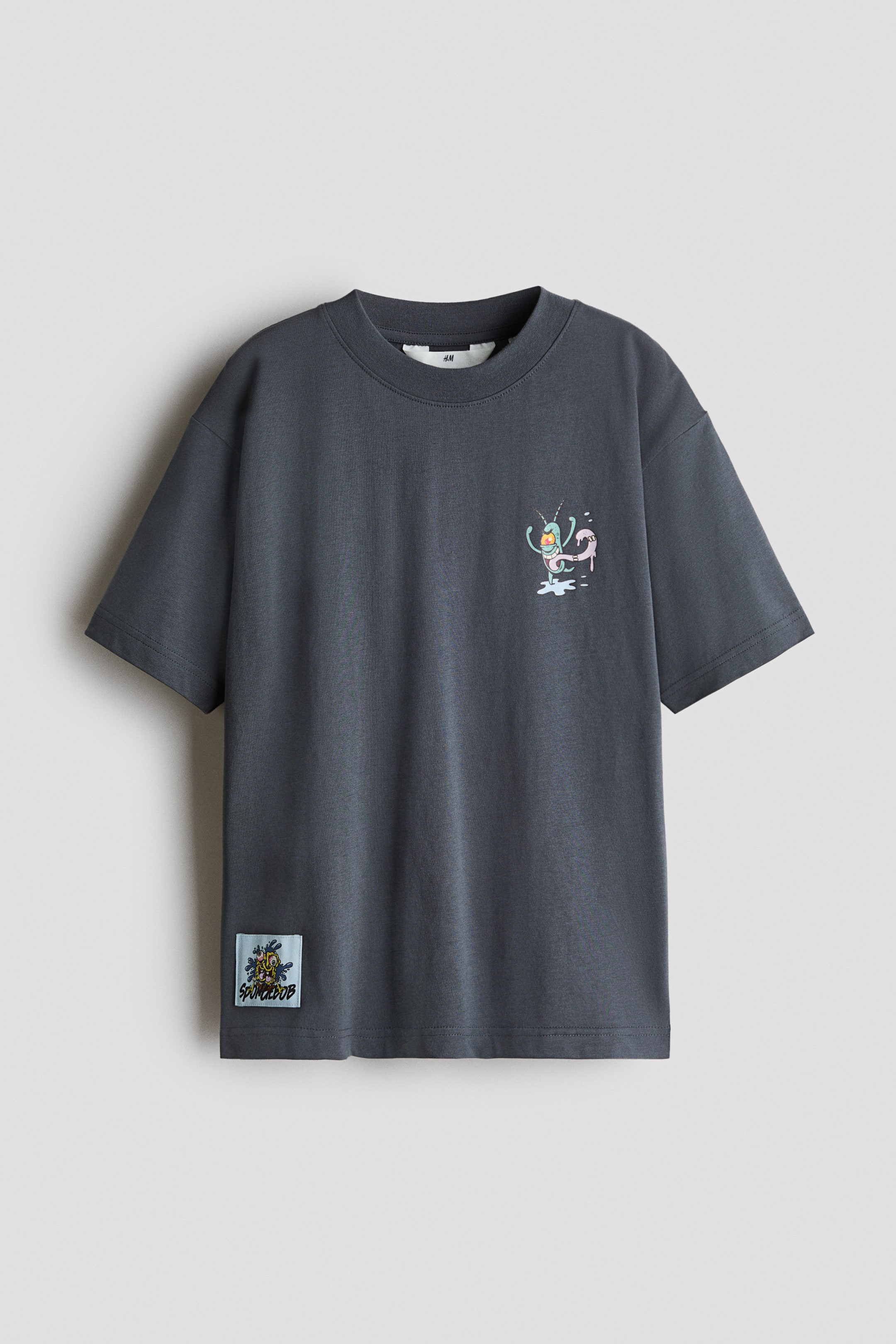 View larger image: A dark gray cotton jersey T-shirt is shown from the front, featuring a round, rib-trimmed neckline and dropped shoulders. It displays a small printed cartoon design on the chest and a light blue rectangular patch near the bottom hem.