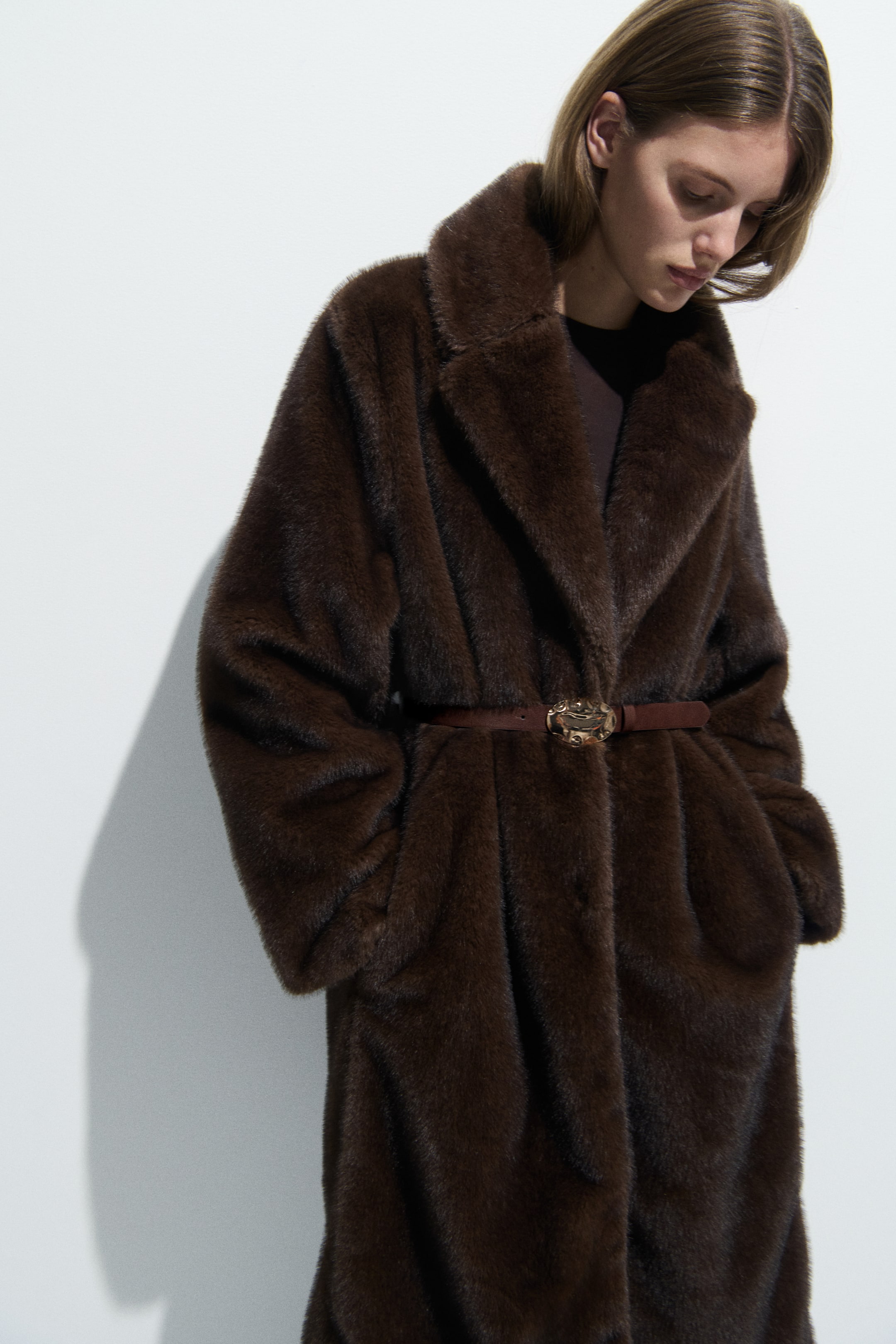 View larger image: A front-facing, dark brown faux fur coat features wide notch lapels, long sleeves, and side pockets. A thin brown belt with a decorative gold-toned buckle cinches the waist.