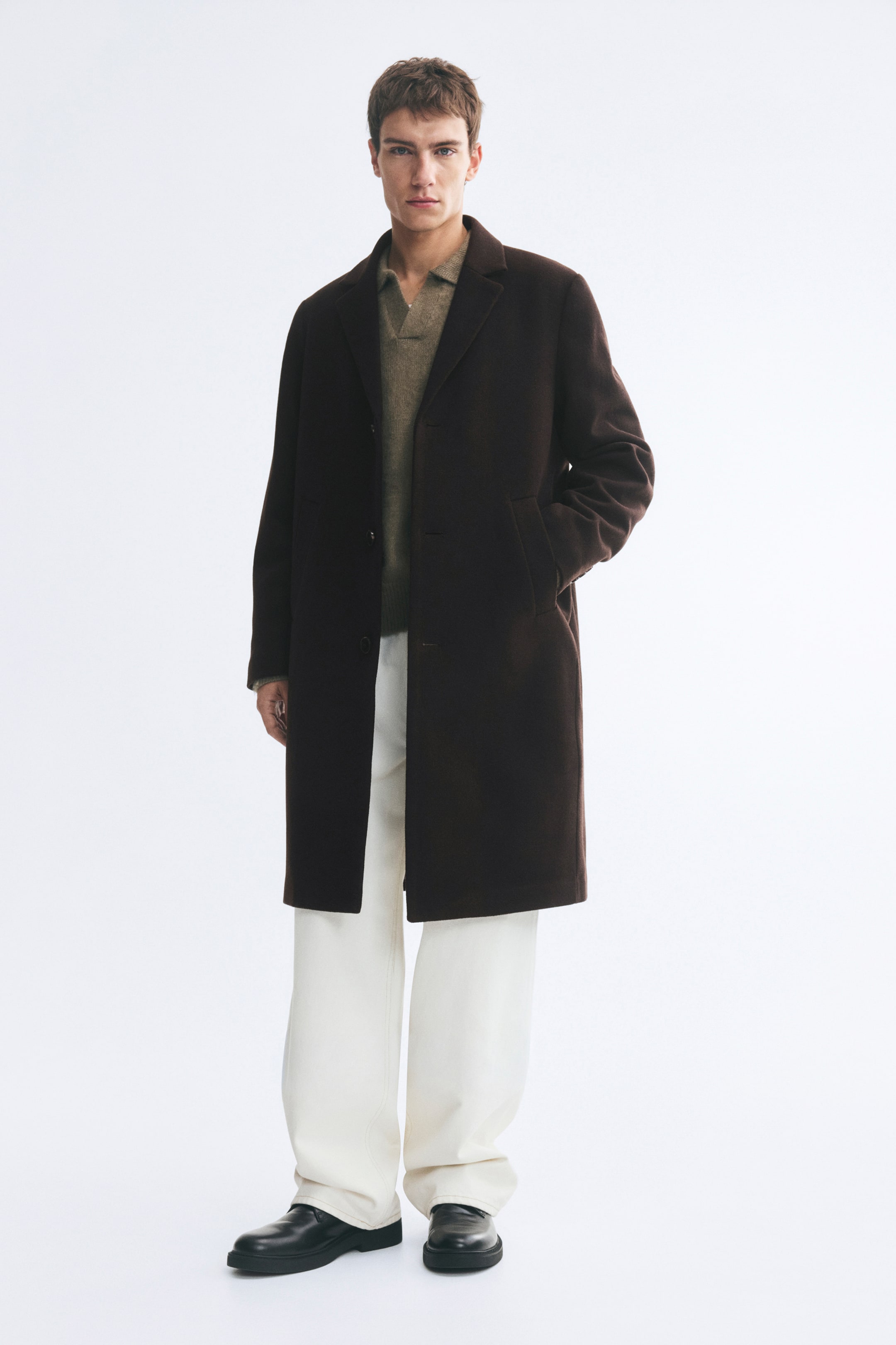 View larger image: A dark brown, knee-length, single-breasted coat with notch lapels and welt pockets is worn by the model. The coat is layered over a brown knit polo sweater and off-white trousers, with the model resting one hand in the side pocket.