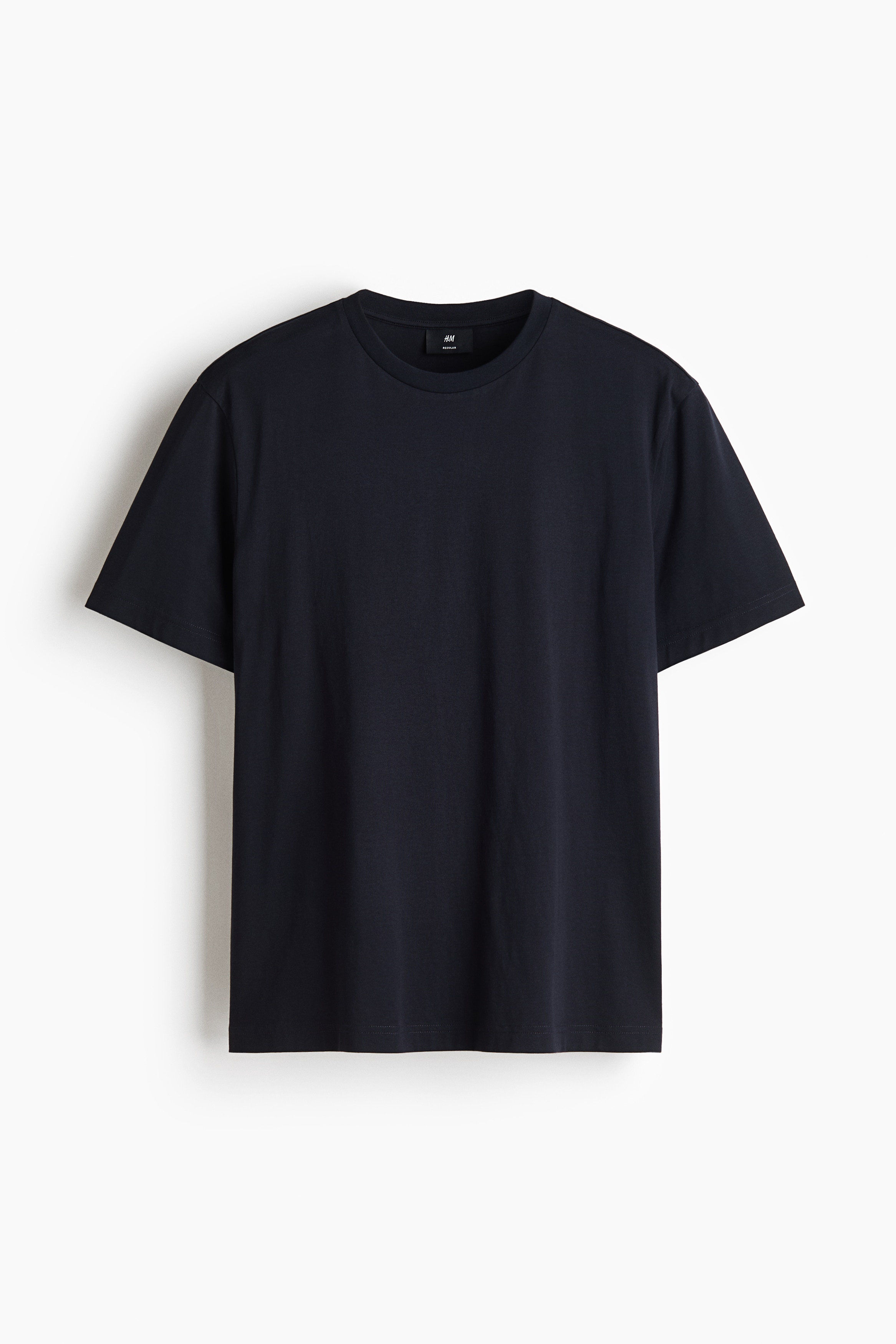 Ein dunkel marineblaues T-Shirt mit kurzen Ãrmeln und Rundhalsausschnitt wird vom Model getragen, das mit den HÃ¤nden in den Taschen einer beigefarbenen Hose steht. Das T-Shirt verfÃ¼gt Ã¼ber eine lockere Passform und eine glatte, klare Textur.