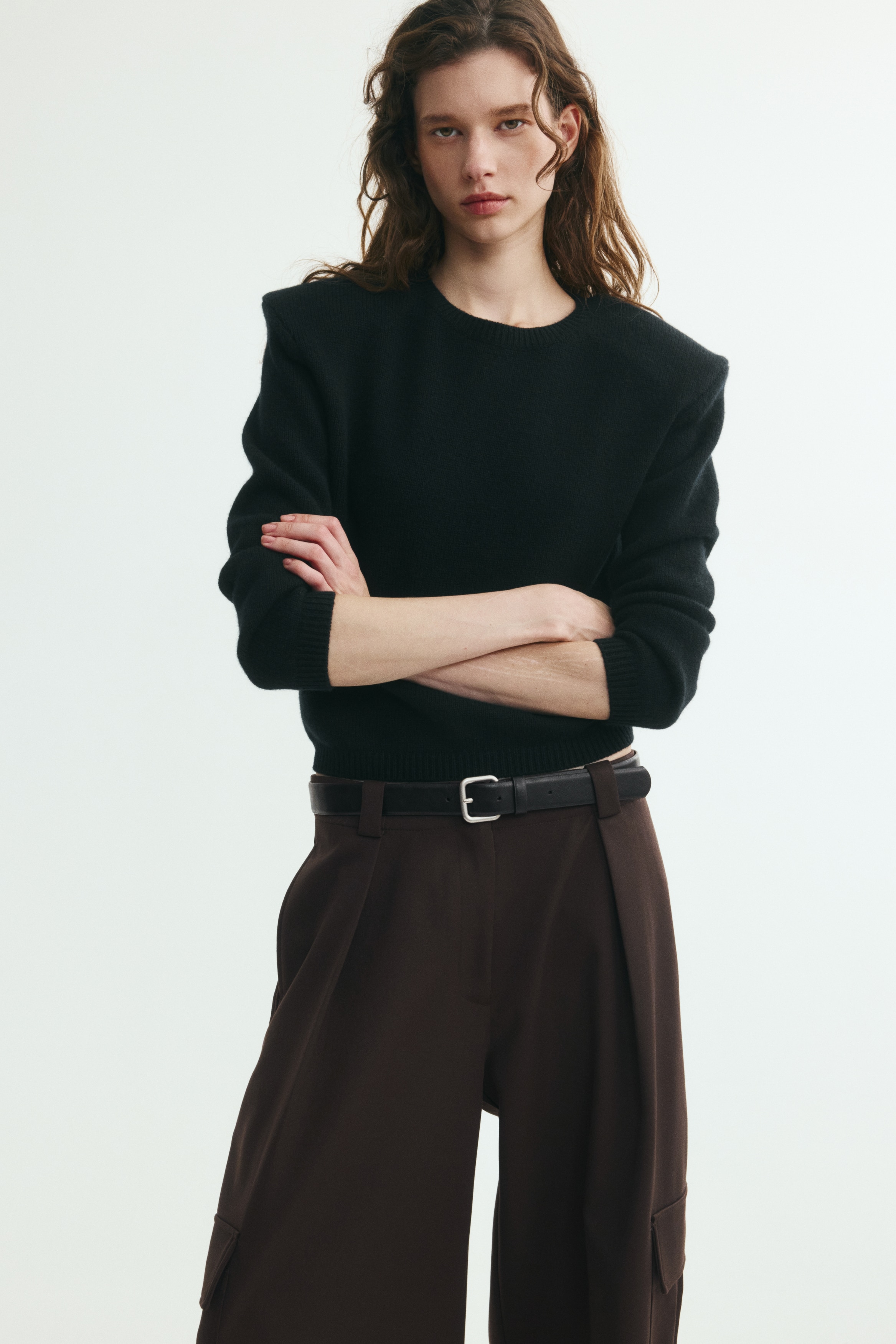 This pair of dark brown, wide-leg trousers is shown from the front, featuring a high waist styled with a slim black belt with a silver buckle. The trousers feature front pleats, side cargo pockets with flaps, and small vertical slit details at the front of the hems.