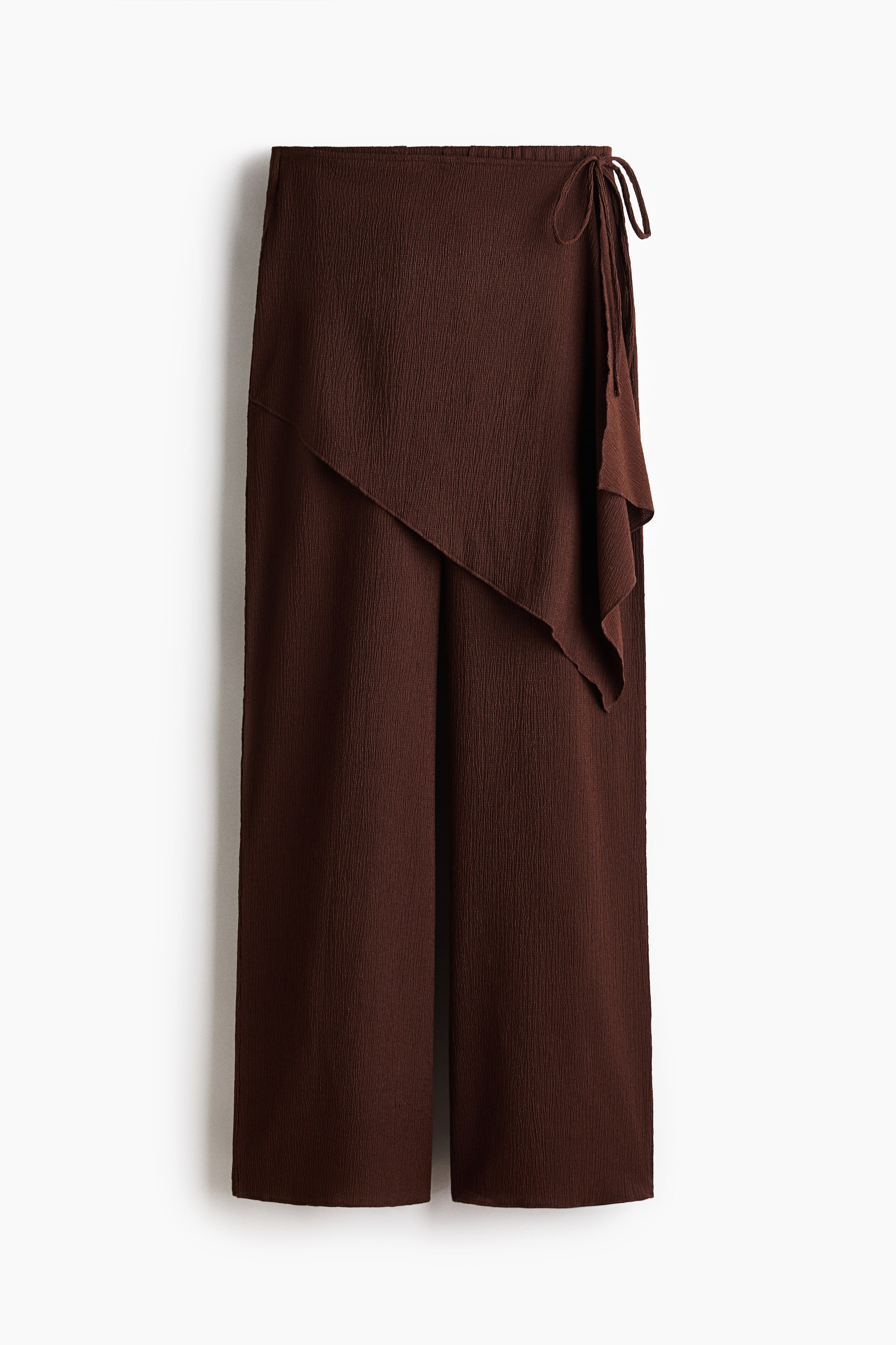View larger image: Dark brown wide-leg pants are presented from the front, featuring a crinkled texture and an asymmetrical wrap-style overlay that ties at the right waist.