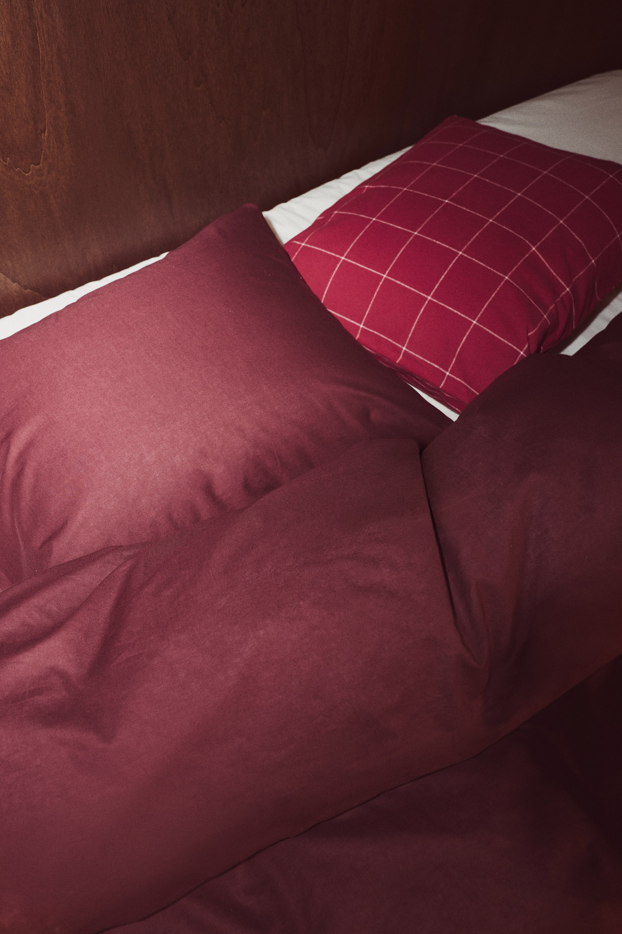 View larger image: A single-size quilt cover set in a rich burgundy shade is shown with a matching solid pillowcase and a softly draped quilt cover. Viewed from a high angle, the set rests on a white bedsheet alongside a styled red-and-white grid pillow.