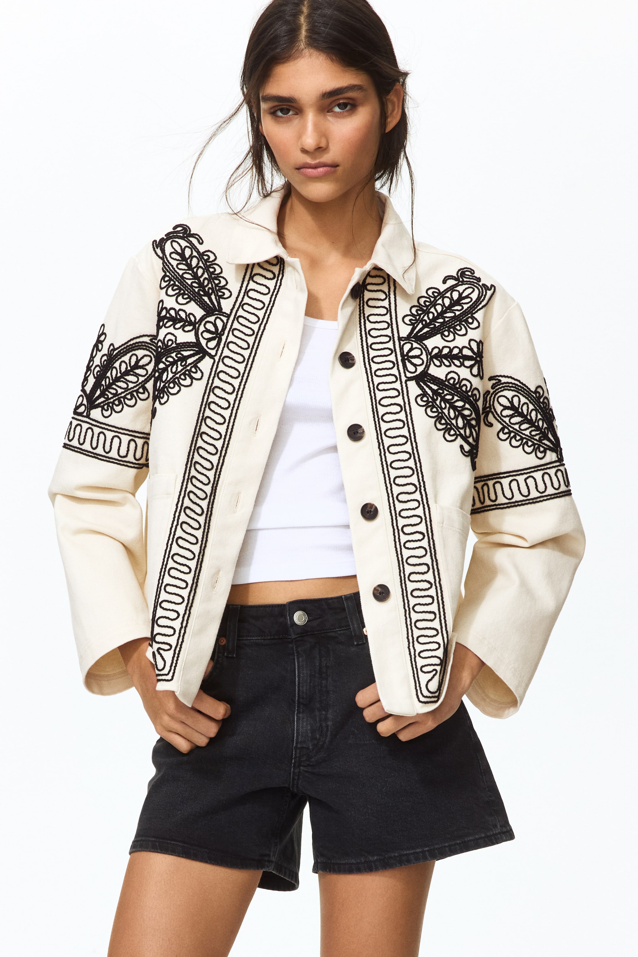 View larger image: An off-white jacket showcases intricate black floral and leaf embroidery on the front panels and sleeves, with a wavy embroidered border accentuating the front placket. It features a pointed collar and dark buttons, styled open to reveal a white tank top underneath.