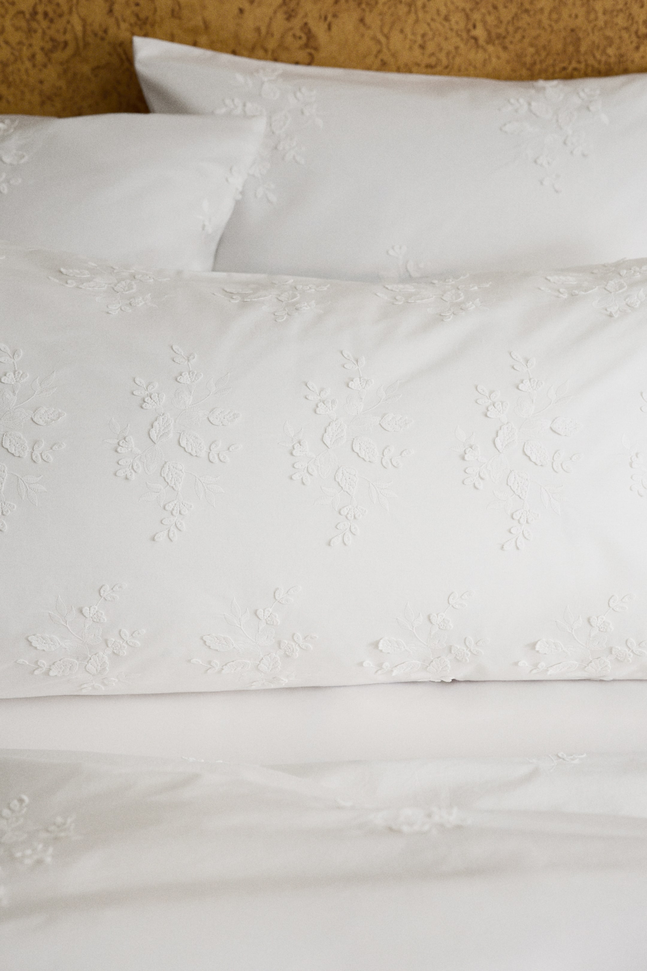 Ver imagen más grande: Una funda de almohada blanca muestra un tejido suave de algodón con intrincados patrones florales bordados en blanco que cubren toda su superficie frontal. Presenta una abertura tipo sobre en la parte trasera, y detrás se puede ver una funda de almohada a juego.