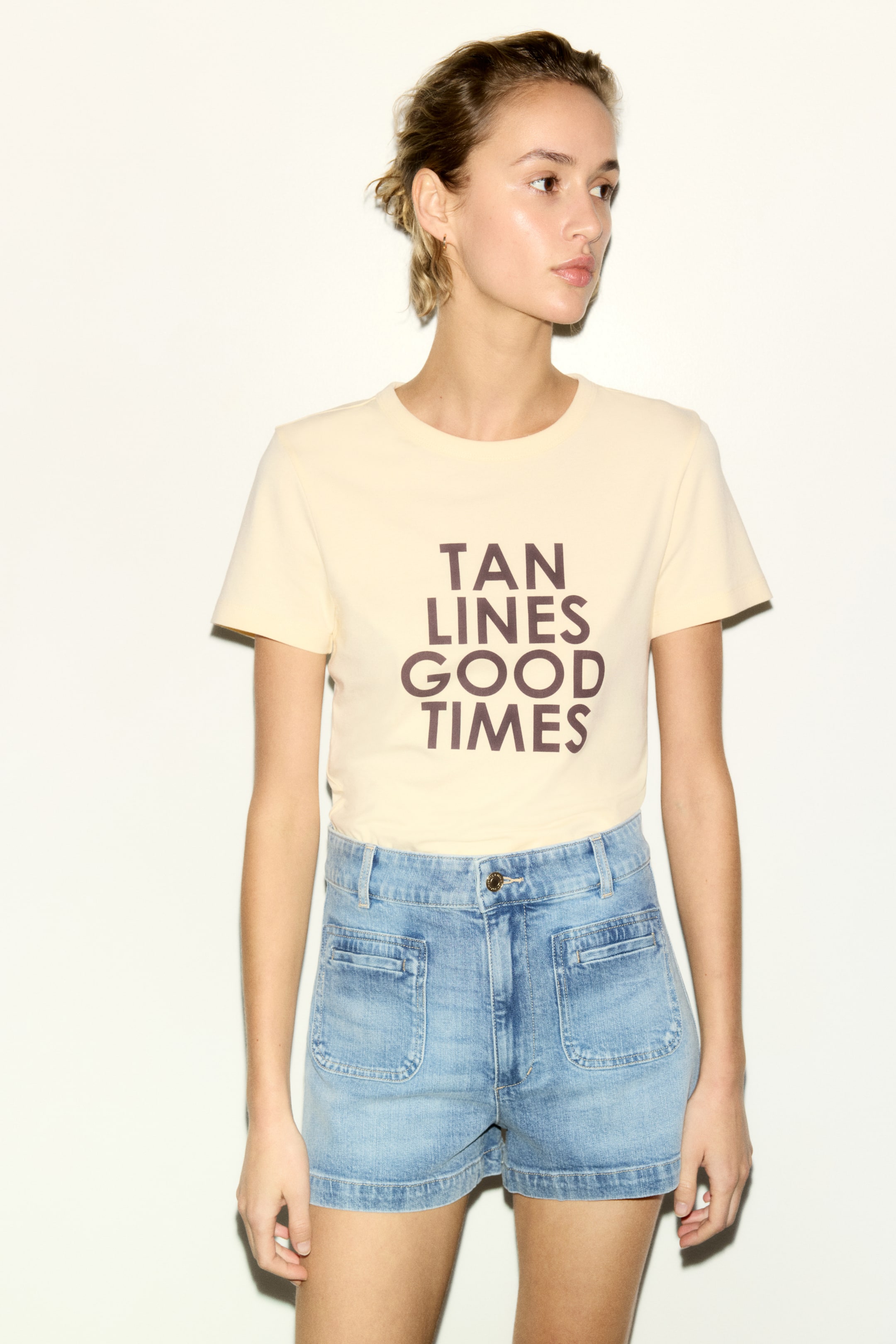 Se større billede: En lysegul t-shirt med rund hals har teksten 'TAN LINES GOOD TIMES' i mørkebrun på forsiden og er kombineret med lyse denimshorts. Shortsene er designet med høj talje og fremtrædende forlommer.