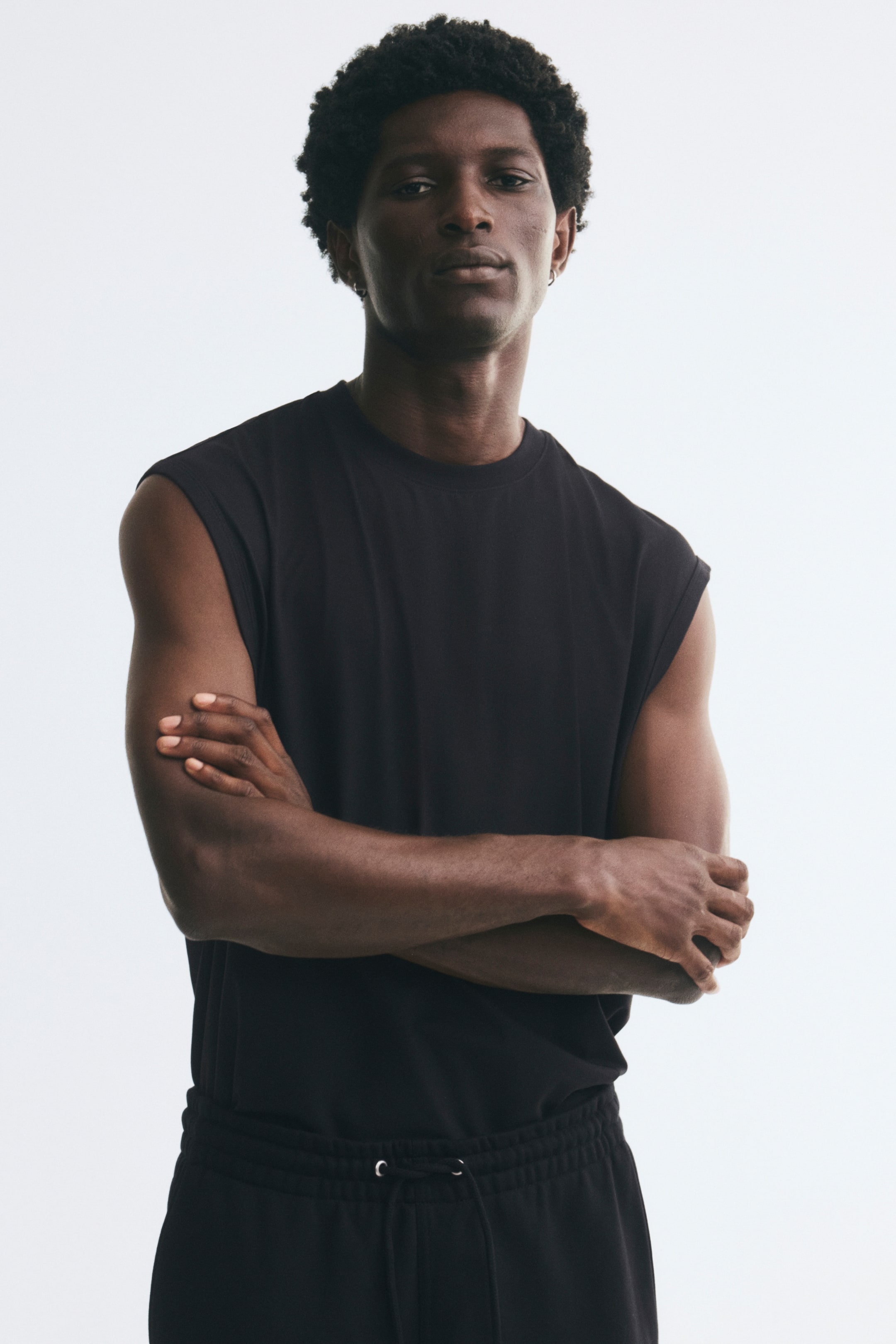 Voir une image plus grande: Un camisole en jersey noir à coupe décontractée, doté d’un col rond bordé de bord-côte et de profondes emmanchures, est porté par le mannequin. Le mannequin est debout, les bras croisés, avec le camisole rentré dans un pantalon de survêtement noir assorti à cordon de serrage.