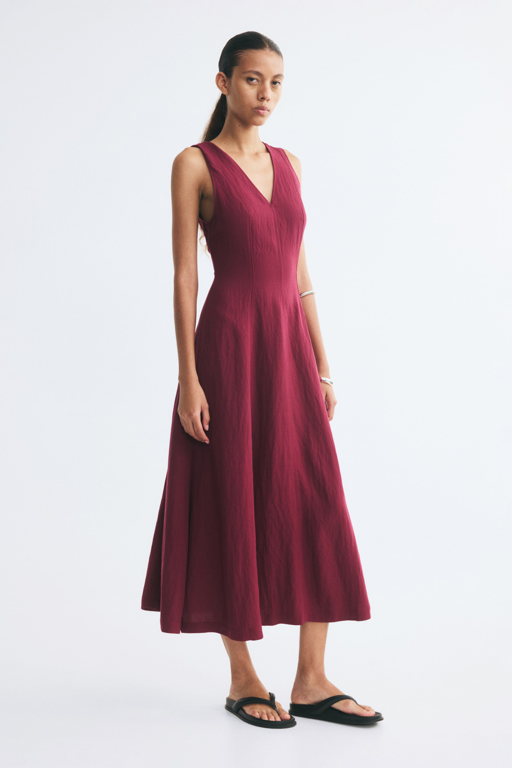 View larger image: A burgundy, sleeveless, calf-length dress is being worn by the model, featuring a V-neckline, vertical seams on a fitted bodice, and a flared skirt. The dress is styled with black flat thong sandals.