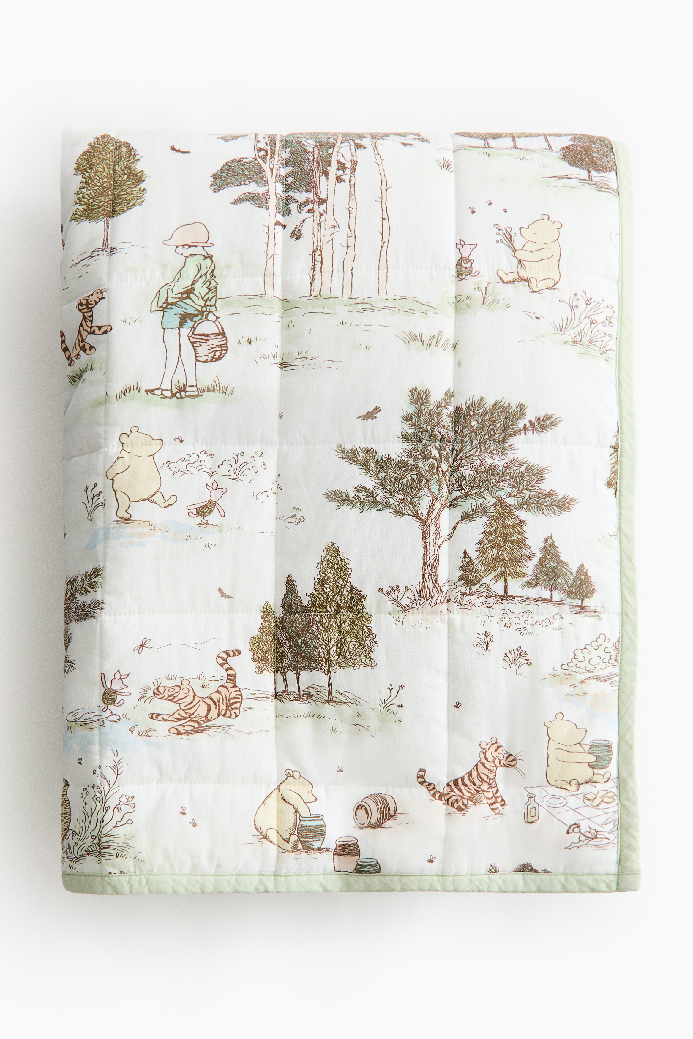 A quilted blanket is lying flat on a bed, displaying a grid pattern and an illustrated scenic print featuring forest elements and figures like Winnie the Pooh, Tigger, and Christopher Robin. It is resting on a light-colored bed frame with a curved side rail, viewed from a slight angle.