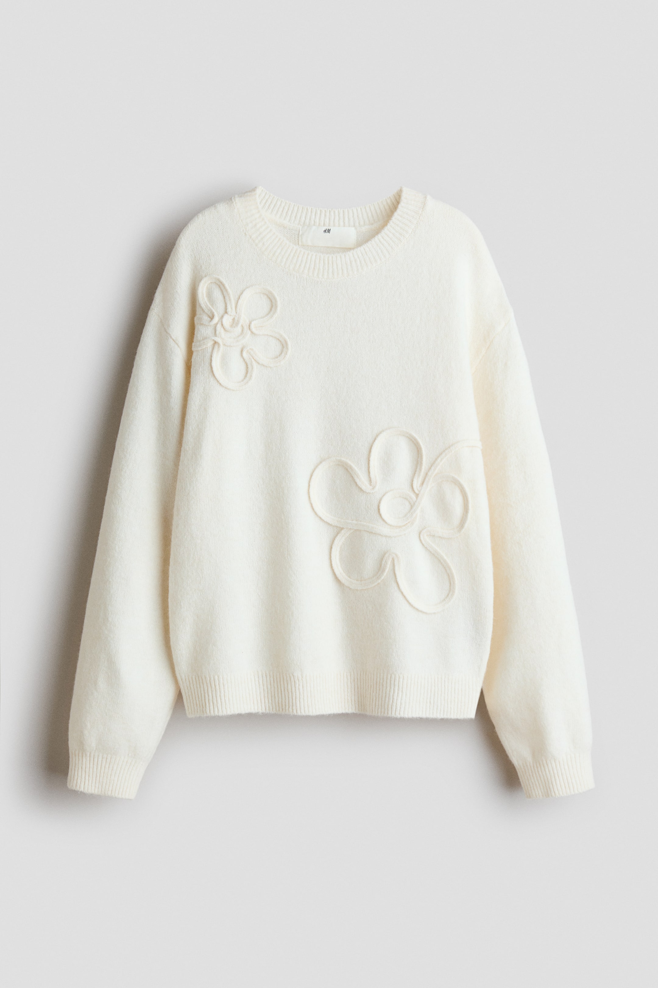 View larger image: The cream sweater is presented FRONT-facing, displaying a ribbed crew neck, cuffs, and hem. Two large outline flower appliqués are adorning the garment, positioned on the upper left and lower right.