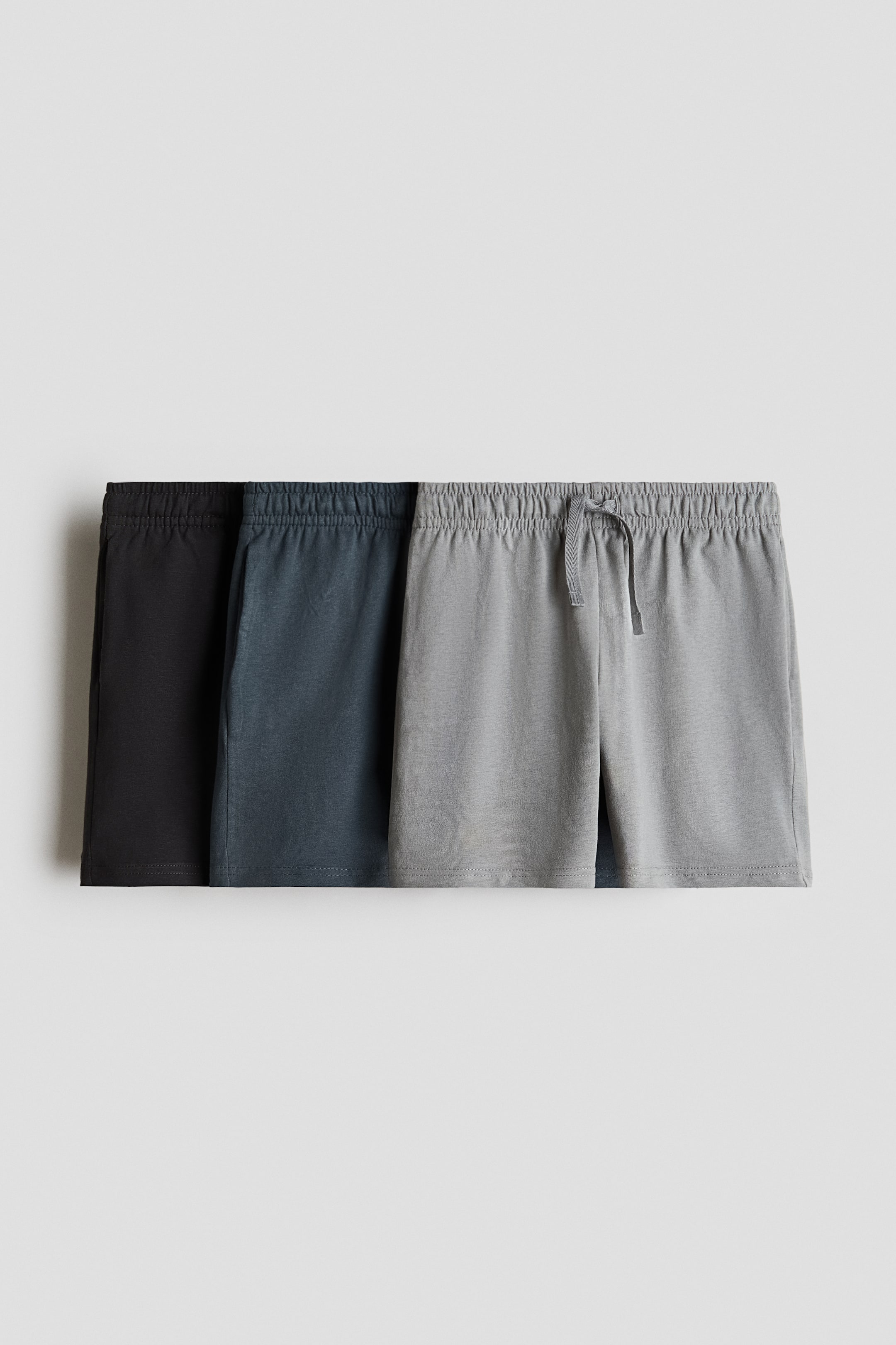 View larger image: "Knee-length shorts crafted from soft cotton jersey are showcased in the foreground, highlighting an elasticated, drawstring waistband and subtle pockets along the side seam. The collection displays three pairs available in black, dark grey, and light grey."