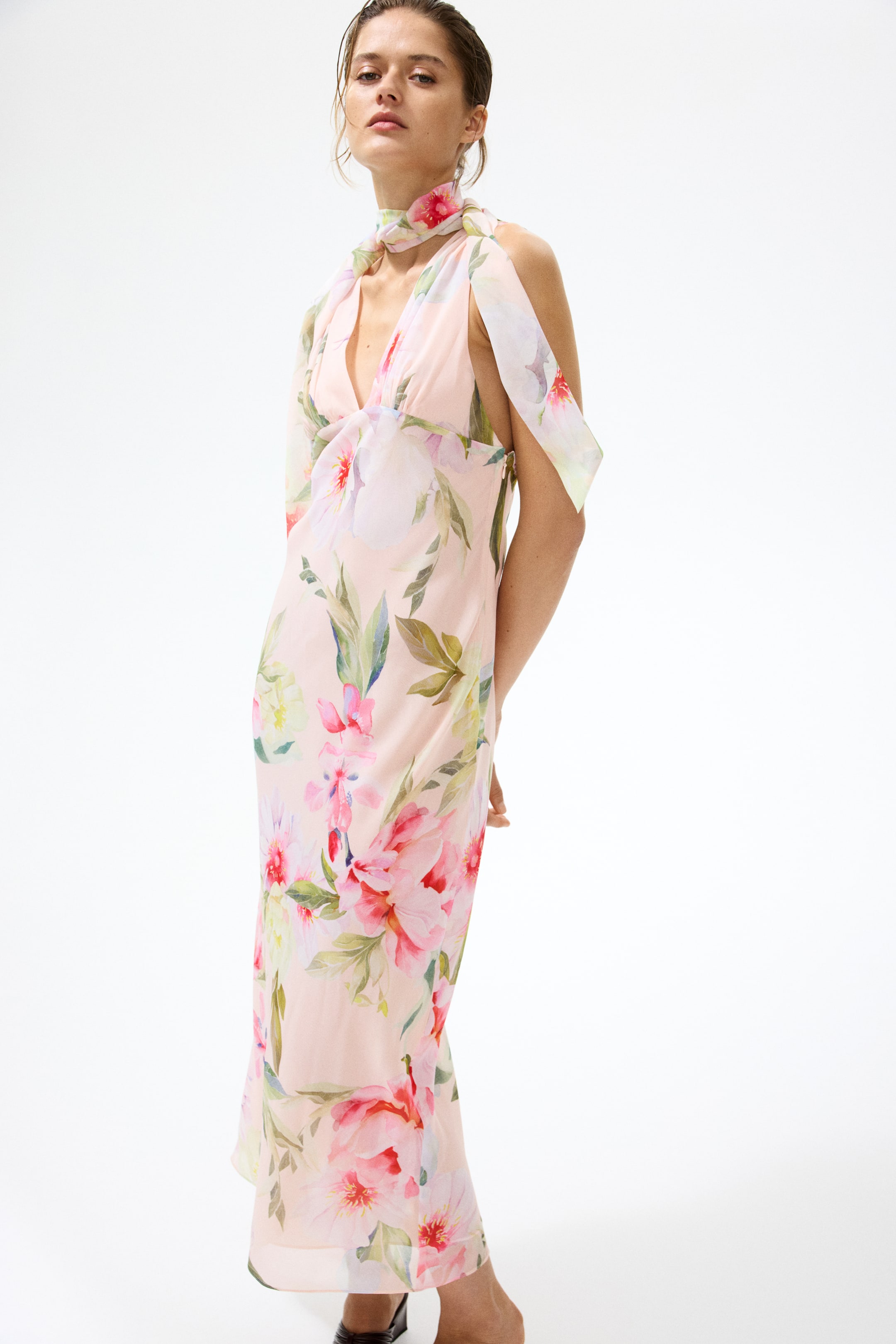 Visualizza immagine più grande: L'abito lungo rosa chiaro presenta una profonda scollatura a V con spalline in stile halter che si estendono e drappeggiano sulle spalle. Mostra una vivace stampa all-over di grandi fiori rosa e rossi con foglie verdi, presentata da un leggero angolo anteriore sinistro.