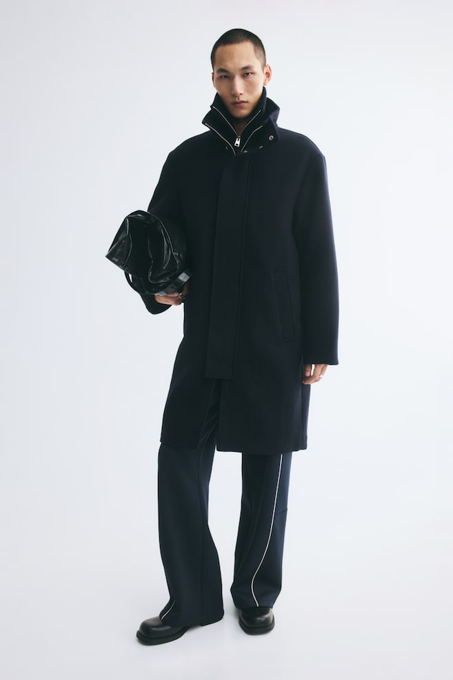 A knee-length, dark navy felted twill coat is being worn by the model, featuring a high funnel collar, a concealed front zip closure with a wind flap, and side welt pockets. The model is styling the coat with dark, side-striped trousers and is carrying a black bag under one arm. - 1