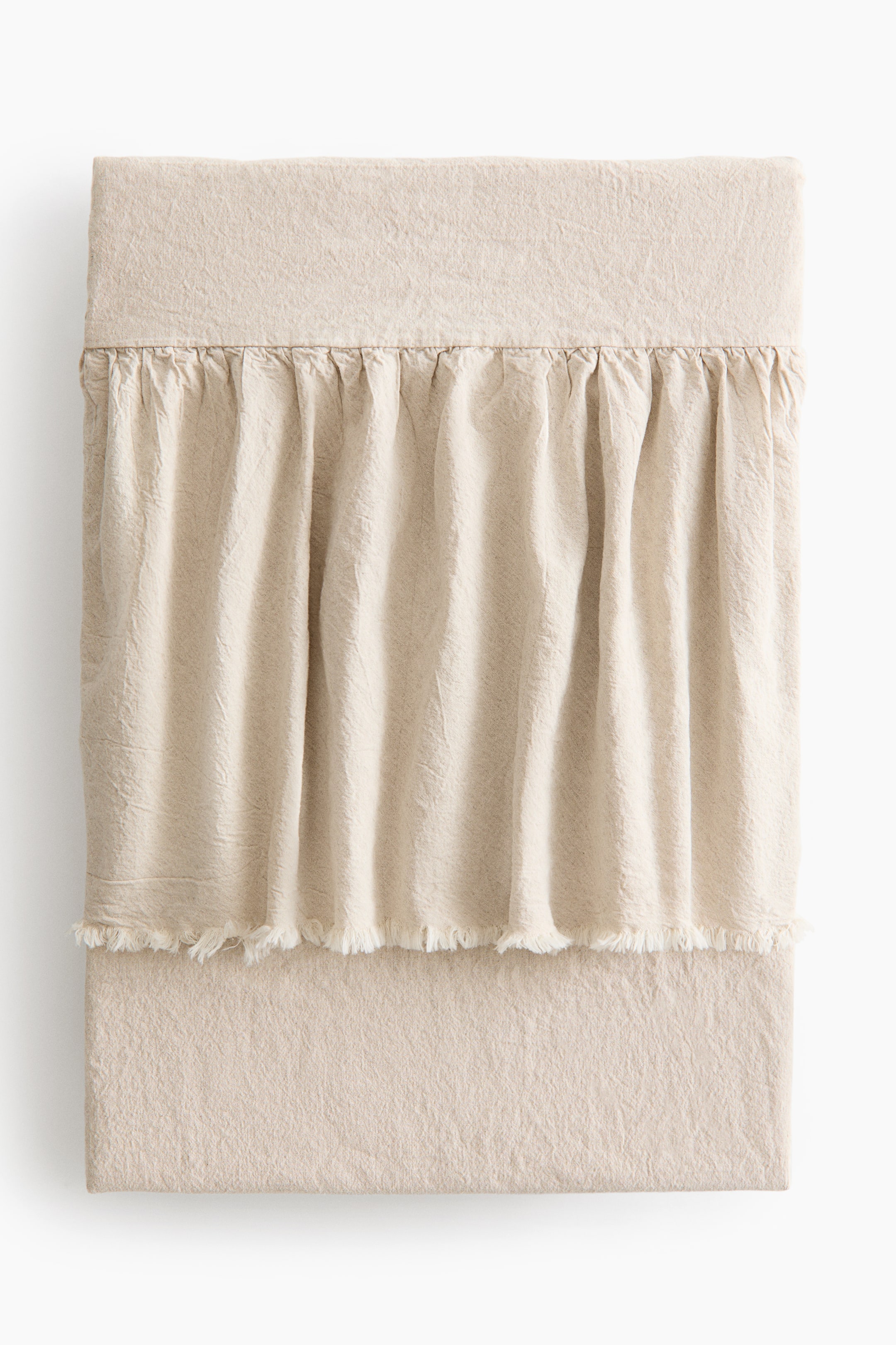 拡大画像を表示: フリルトリムのベッドスプレッドが正面から見え、柔らかくドレープしたライトベージュのウォッシュドリネンとコットンの織りを特徴としています。上部中央には、未加工のフリンジエッジを持つギャザーフリルが水平に走っています。