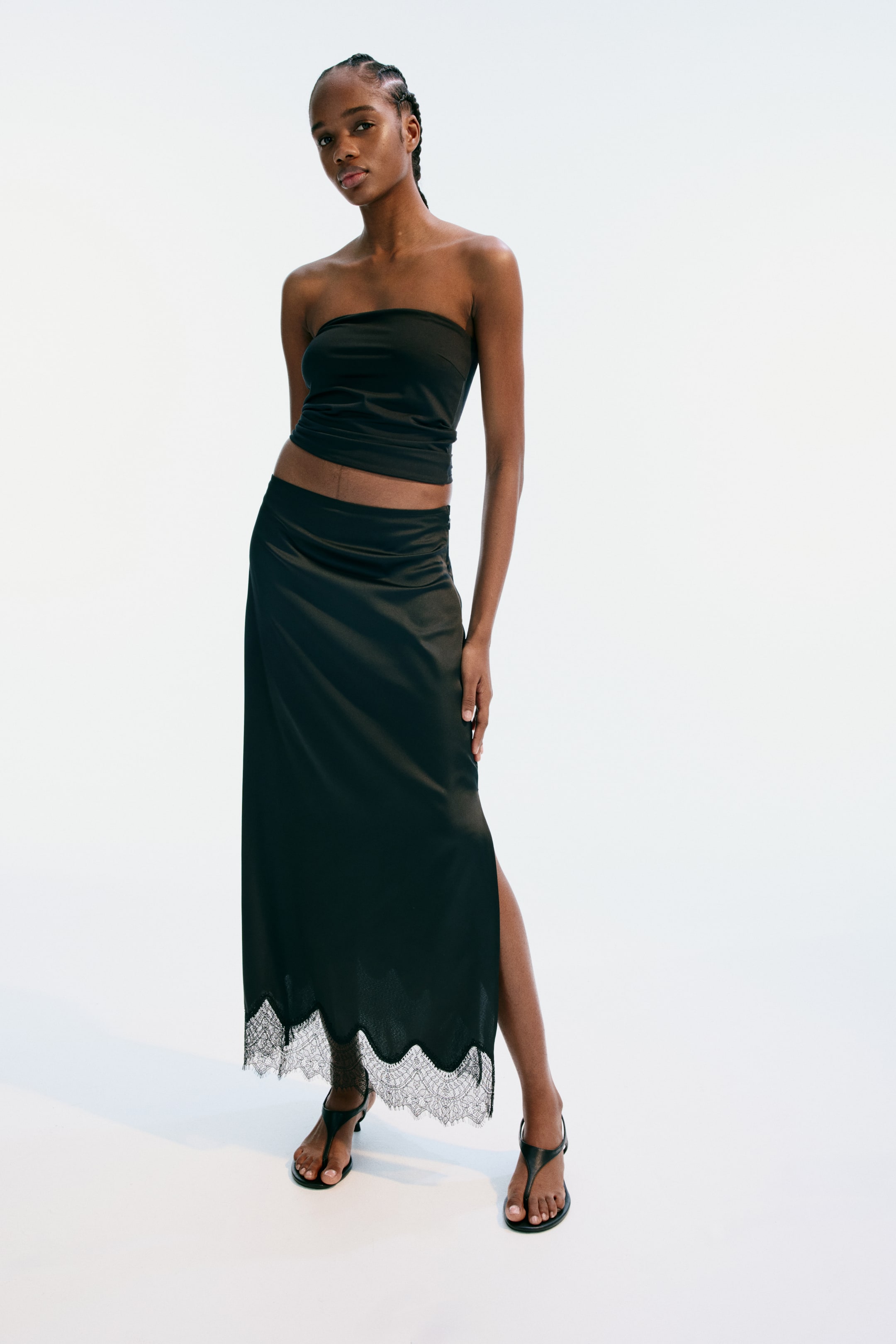 View larger image: A black bandeau top showcases a strapless, cropped design. It's paired with a long black skirt featuring a satin-effect fabric, a high side slit, and a scalloped white lace trim along the hem.