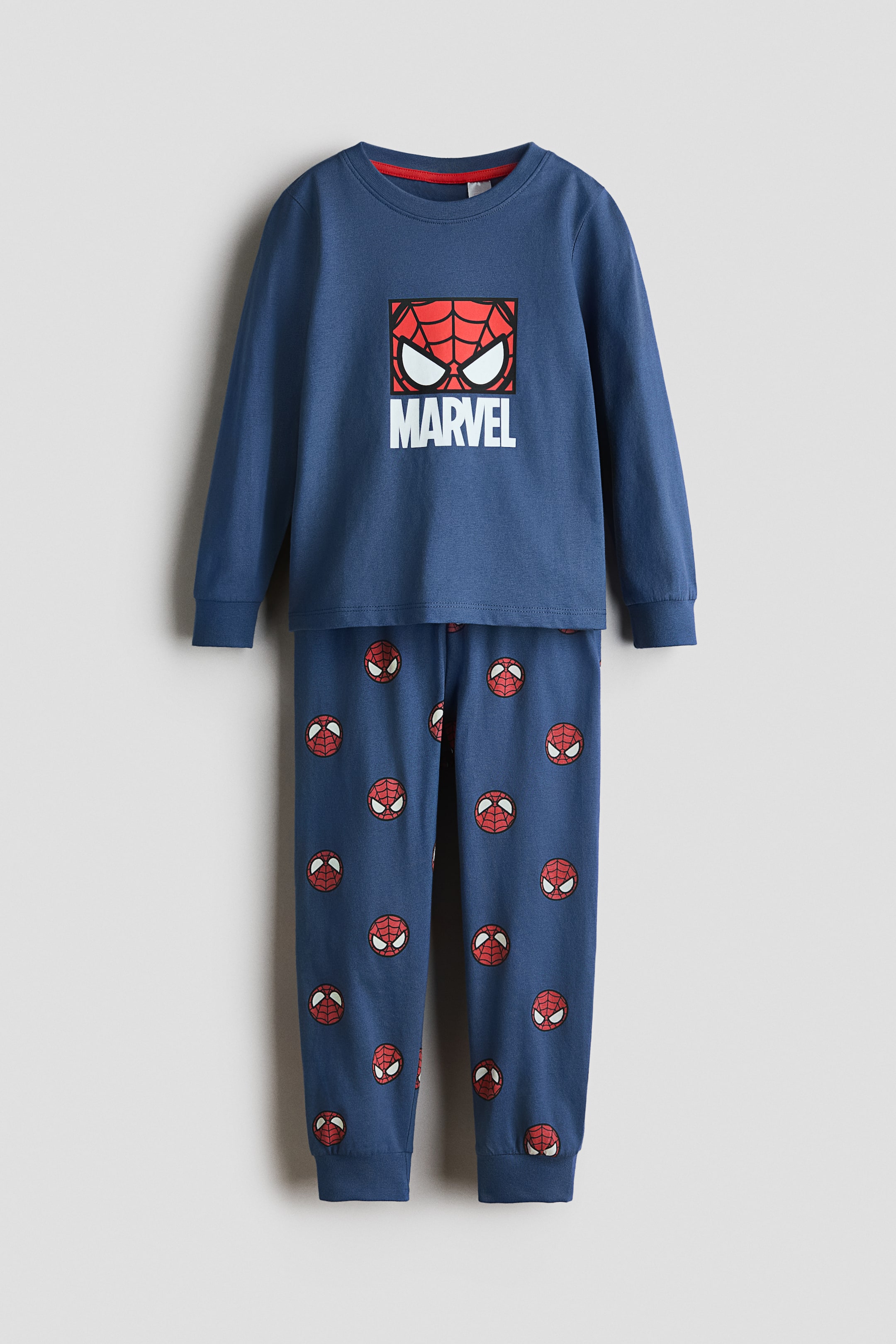 View larger image: A blue two-piece pajama set is showcased from the front, boasting a long-sleeved top with a crew neck, a red inner lining, and a prominent square graphic of a red Spider-Man mask with white eyes and the word 'MARVEL' on the chest. The coordinating long pants display a repeating pattern of small red Spider-Man masks and conclude with elasticated cuffs at the ankles.