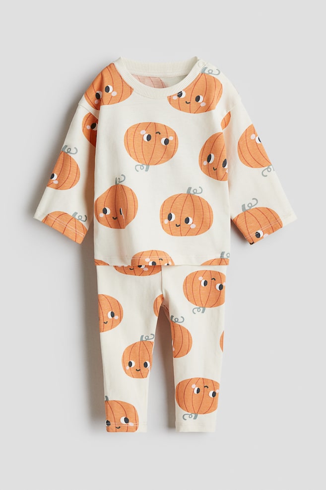 Een tweedelig crèmekleurig babysetje ligt plat en toont een all-over print van oranje, lachende cartoonpompoenen. Het langemouwenshirt heeft een geribbelde ronde hals en drukknoopjes op één schouder, terwijl de lange broek eenvoudige zomen heeft. - 1