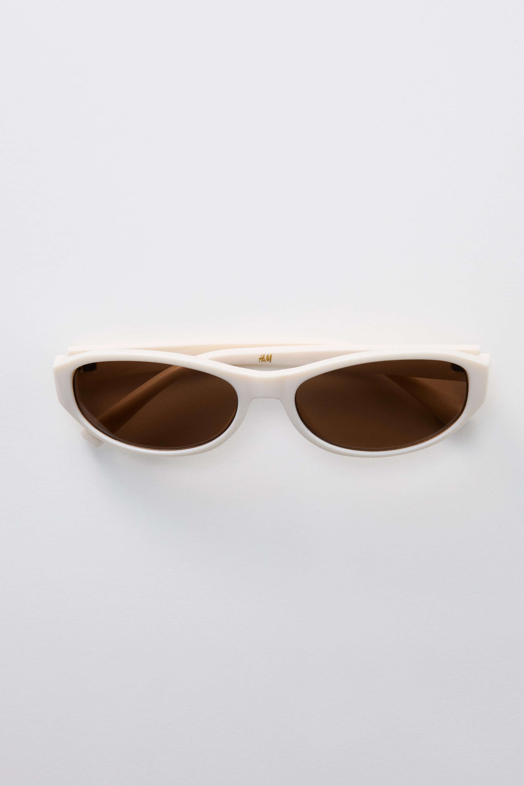 View larger image: Oval sunglasses feature off-white plastic frames and sidepieces. They display tinted brown plastic lenses, and an H&M logo is visible on the right sidepiece.