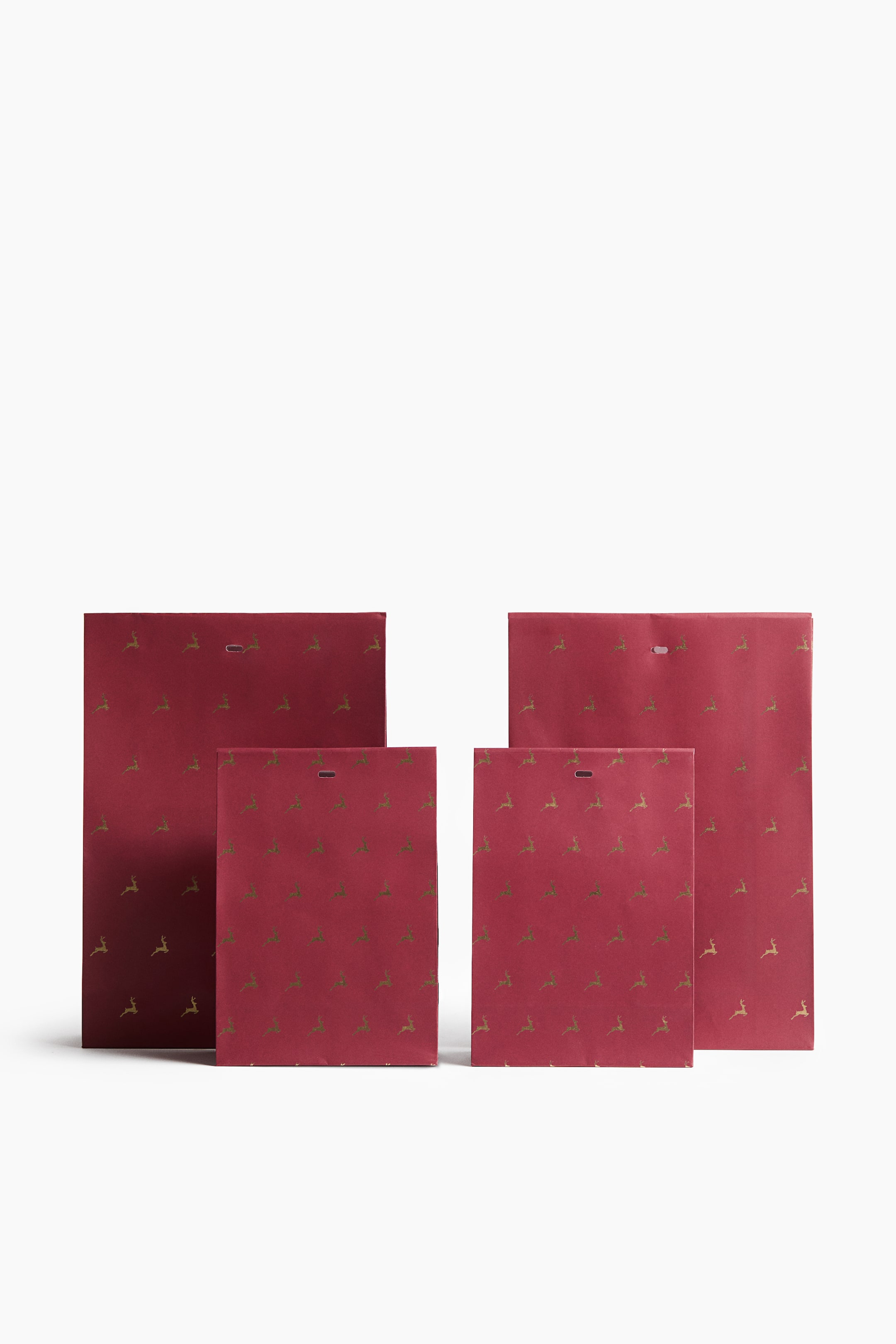 View larger image: A set of four dark red paper gift bags, comprising two large and two small bags, is standing upright facing forward. Each bag features an all-over pattern of small gold reindeer and has a fold-over top flap with a pre-punched horizontal slot.