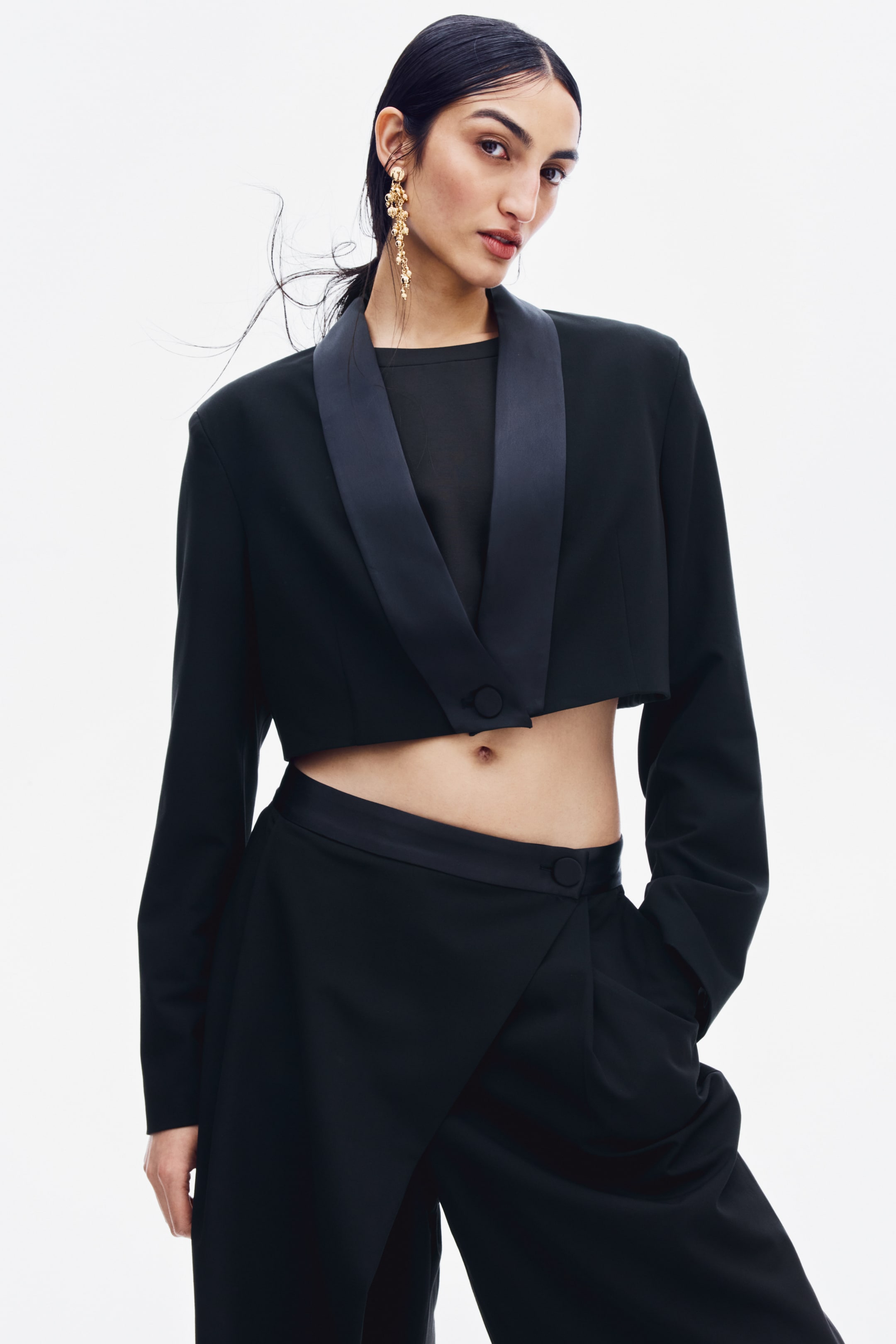 View larger image: A cropped black twill blazer featuring satin shawl lapels and a single covered button is worn by the model. The blazer is layered over a black top and paired with matching black trousers.