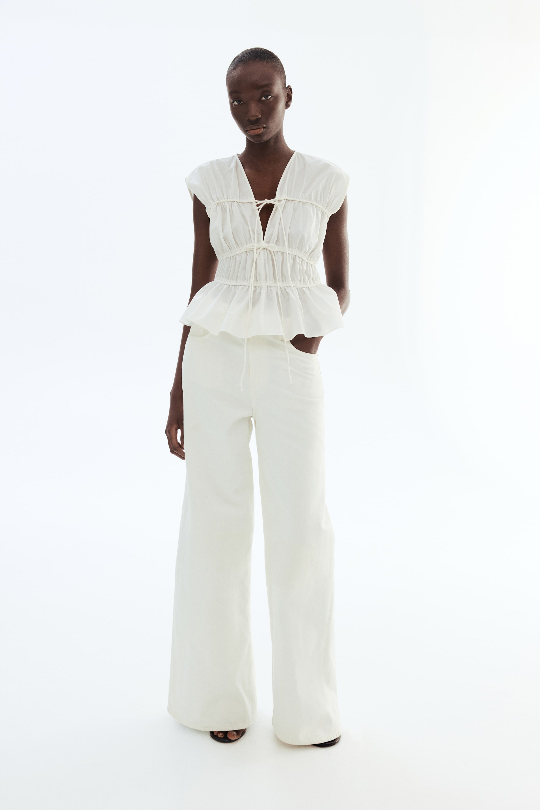 View larger image: A white peplum top paired with matching wide-leg trousers is showcased from a front-facing perspective. The top highlights a lace-up V-neck, a shirred bodice with short cap sleeves, and a flared peplum hem. The trousers exhibit a relaxed fit, featuring visible side pockets and a drawstring waist.