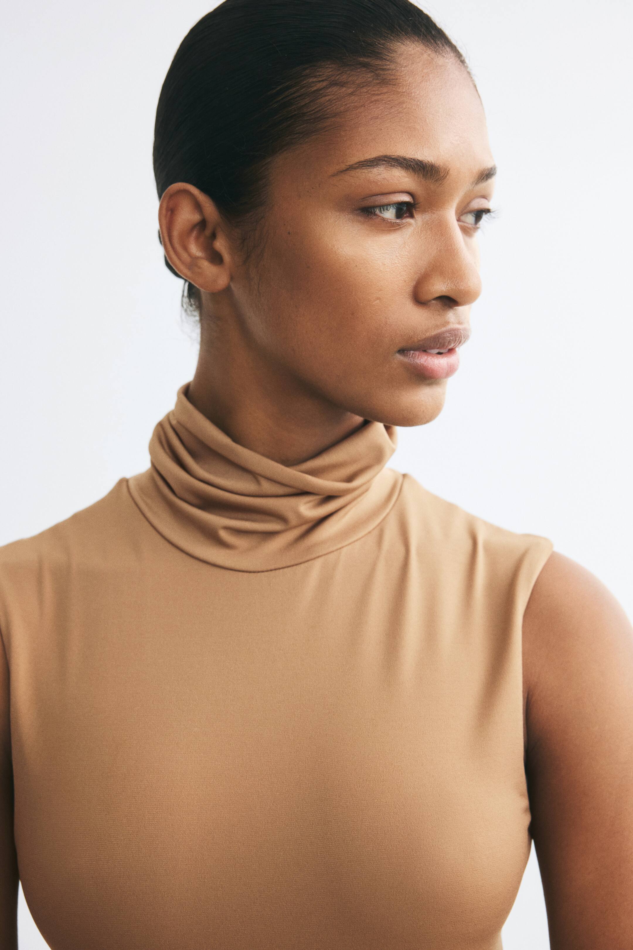 View larger image: A camel-coloured sleeveless top with a draped turtleneck is worn by the model. The fitted top is made from a smooth, stretch fabric.