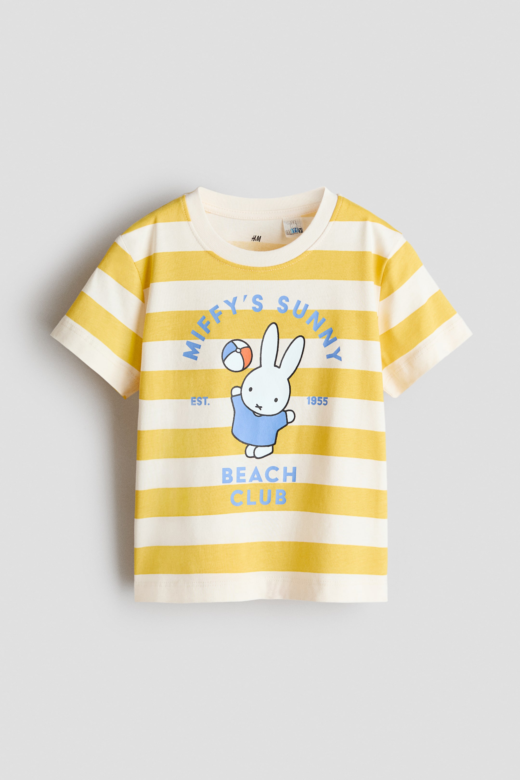 Ampliar la imagen: Una camiseta de frente en suave jersey de algodón muestra rayas horizontales amarillas y crema. Presenta un cuello redondo y mangas cortas, con un motivo impreso en el frente que representa a un personaje de conejo blanco con una camisa azul sosteniendo una pelota de playa, y texto azul que dice "Miffy's Sunny" arriba, "Beach Club" abajo, y "Est. 1955" flanqueando al conejo.