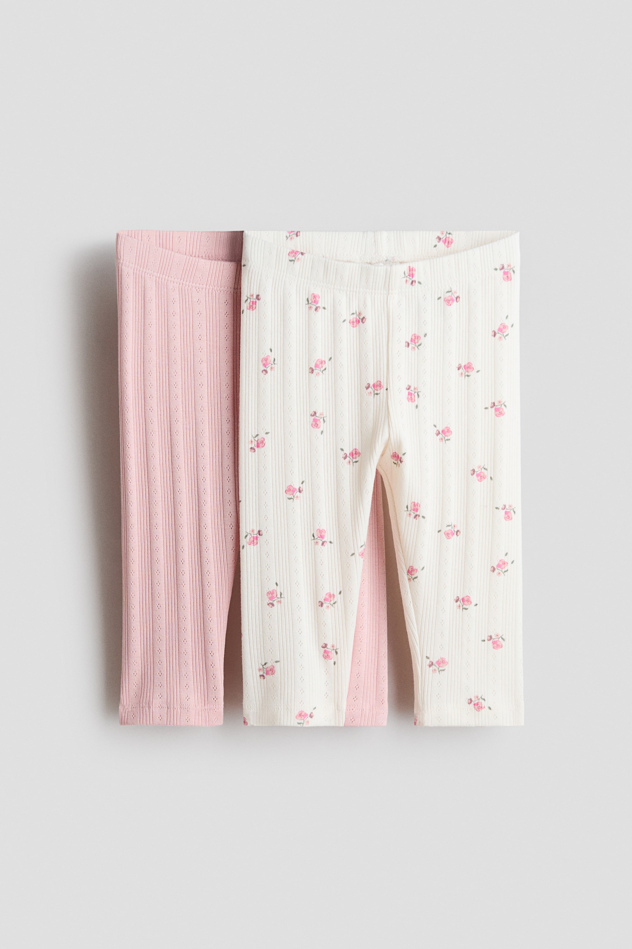 Zobrazit větší obrázek: Legíny jsou zobrazeny zepředu; jde o dva páry z žebrovaného dresu s detaily typu eyelet. Jeden pár je světle růžový, druhý krémový s jemným květinovým potiskem drobných růžových růžiček; oba mají elastický pas.