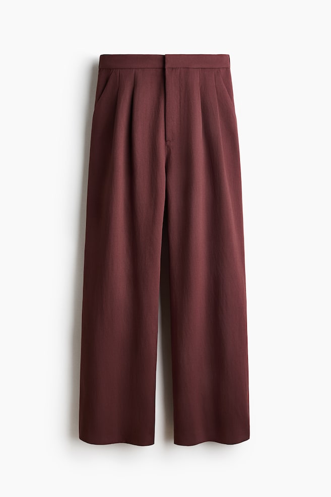 Burgundy wide-leg trousers feature a flat waistband with a visible button fastening above a zip fly. Single pleats extend from the waist, and side slash pockets are visible. - 1