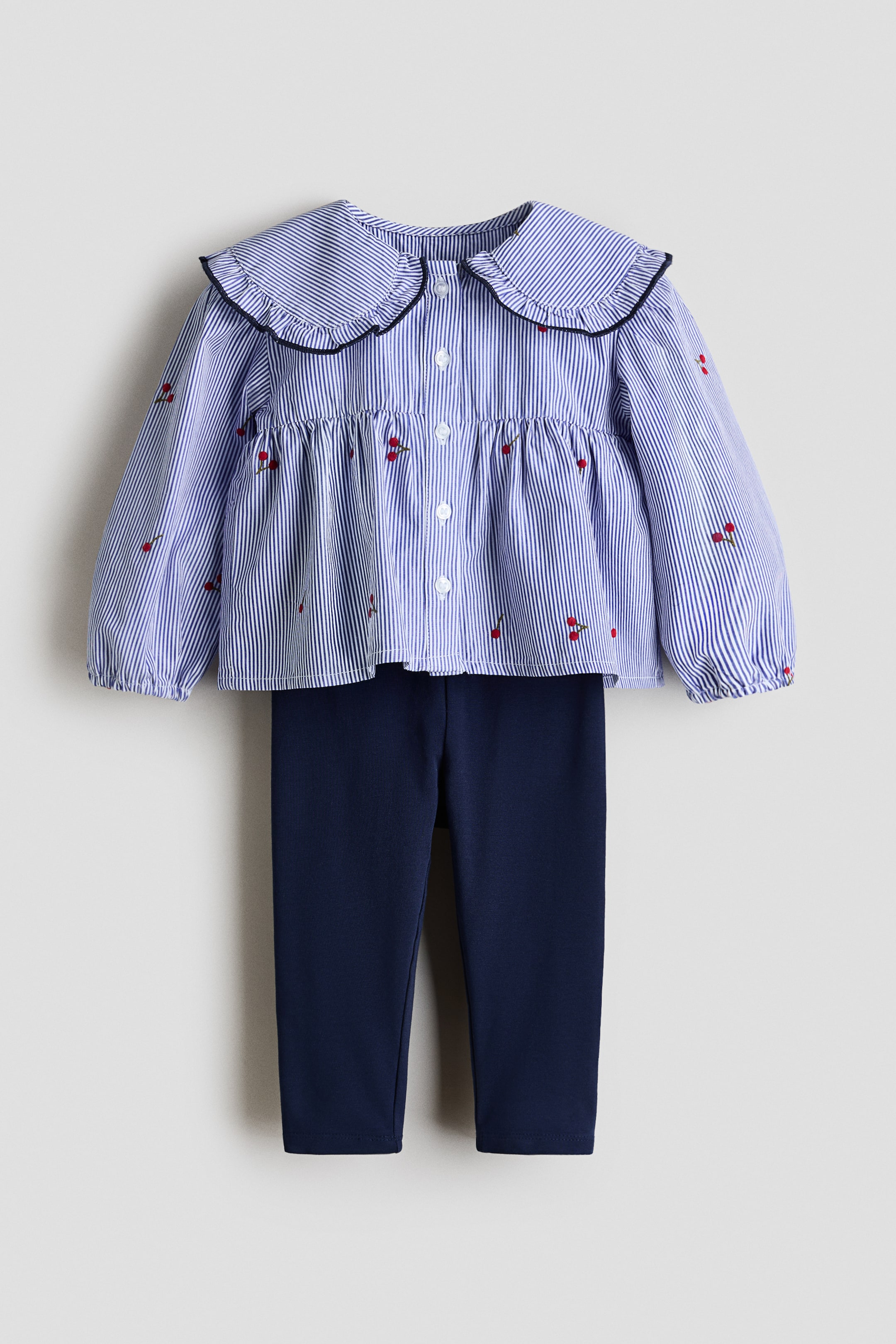 View larger image: A two-piece children's outfit showcasing a blue and white striped blouse along with navy blue leggings, facing front. The blouse features a ruffled Peter Pan collar outlined in black, a button-down front, gathered elasticated cuffs, a gathered peplum hem, and small red cherry embroidery throughout.