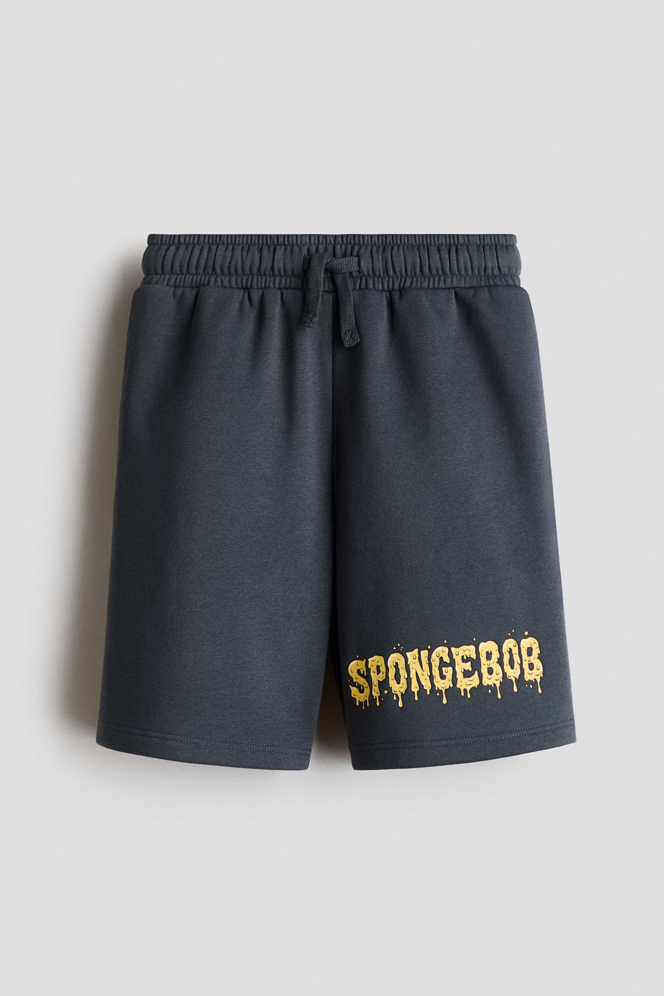 View larger image: Dark grey fleece shorts are shown from the front, featuring an elasticated waistband with a knotted drawstring. The shorts display a yellow, dripping-style graphic print reading "SPONGEBOB" on the lower left leg.