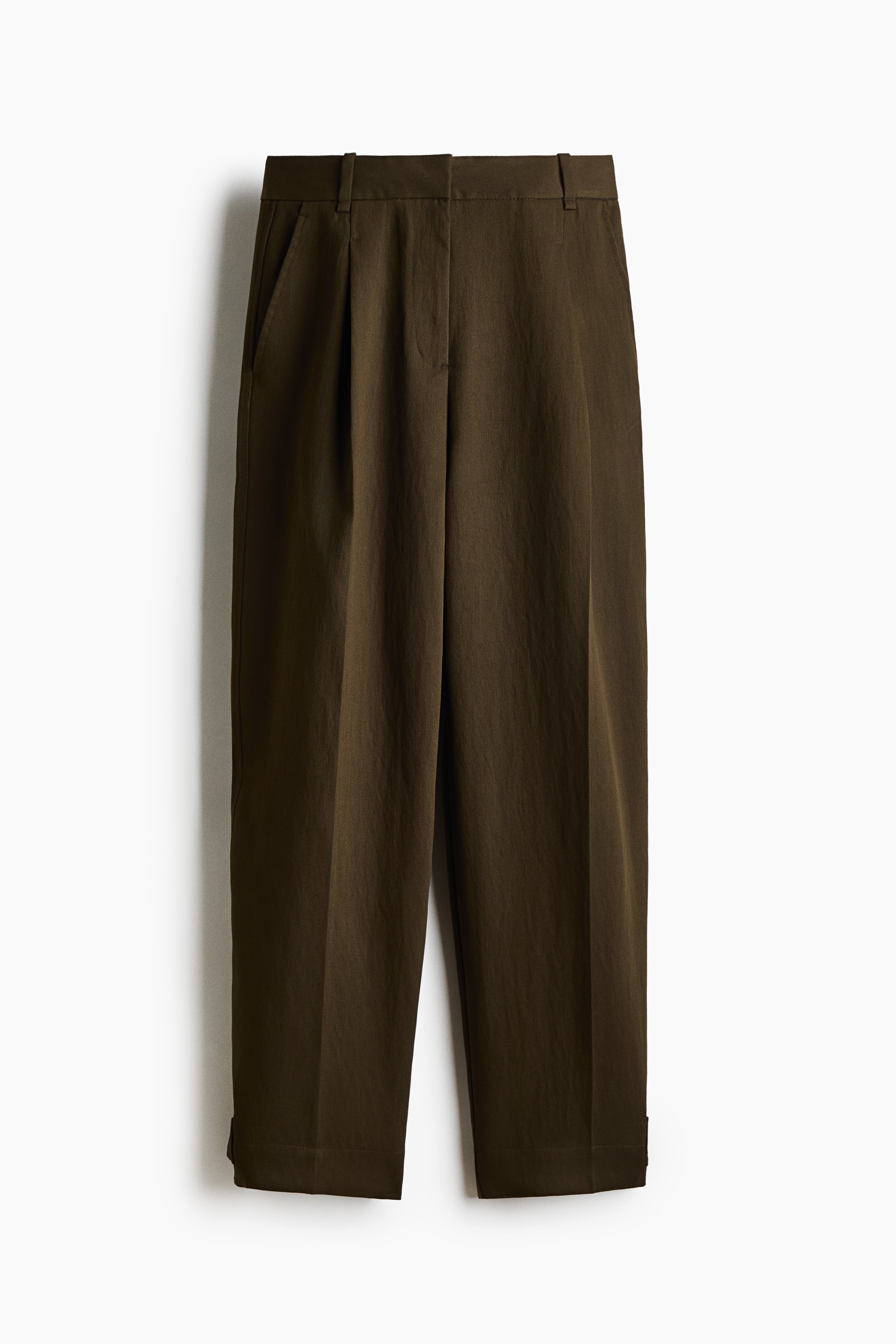 Losvallende donkerbruine broek met hoge taille, voorzien van gestructureerde taillebandflappen, plooien aan de voorkant en geperste vouwen, gedragen door het model. Het model staat terwijl de broek wordt gestyled met een zwart leren jack met hoge kraag.