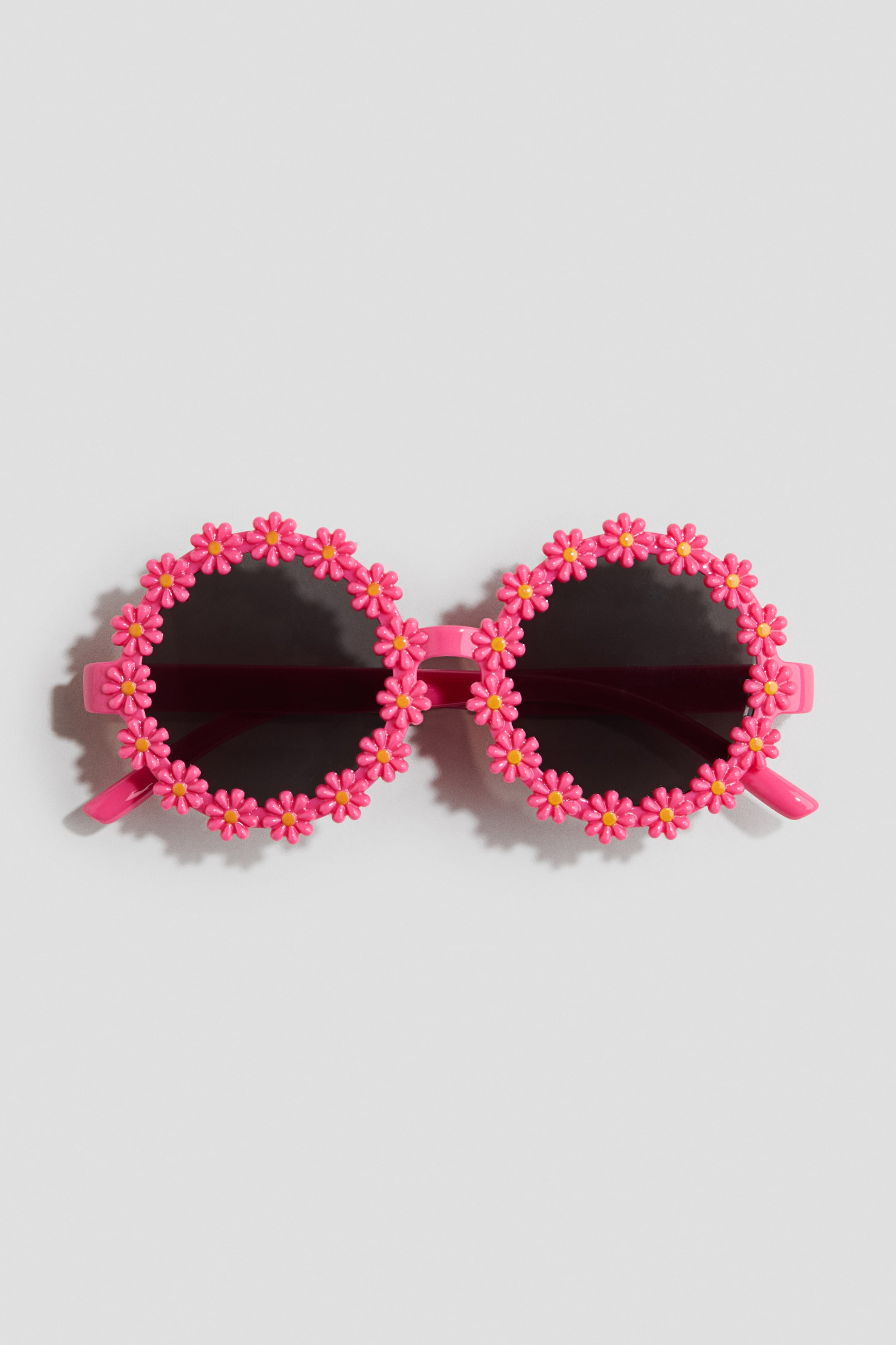 View larger image: These sunglasses showcase a frontal perspective, featuring round, dark lenses encased in vibrant pink frames. Delicate pink flowers with yellow centers embellish the edges of both frames, while pink arms extend gracefully from the sides.