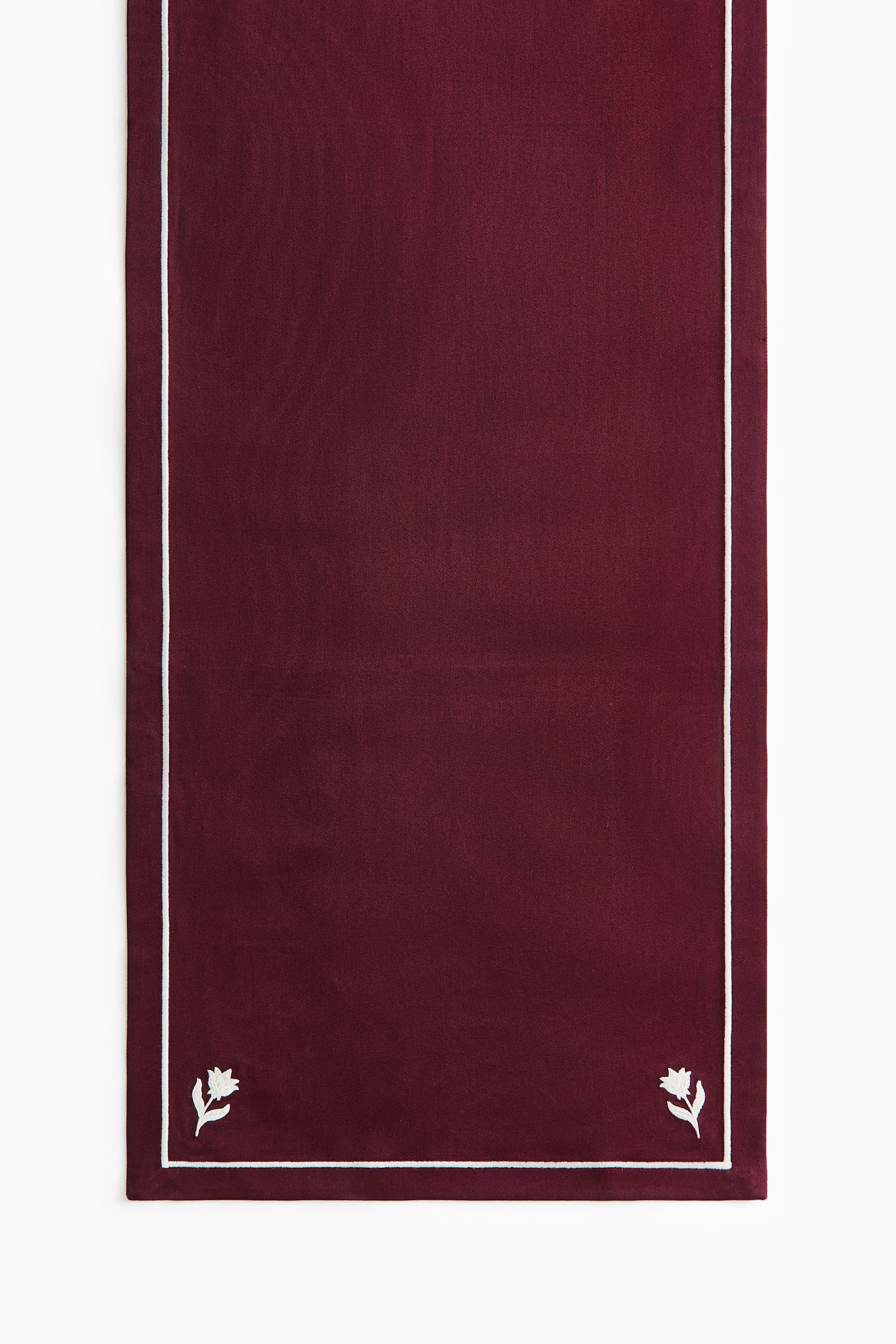 A burgundy table runner features a thin white embroidered border and small white floral motifs in the bottom corners.