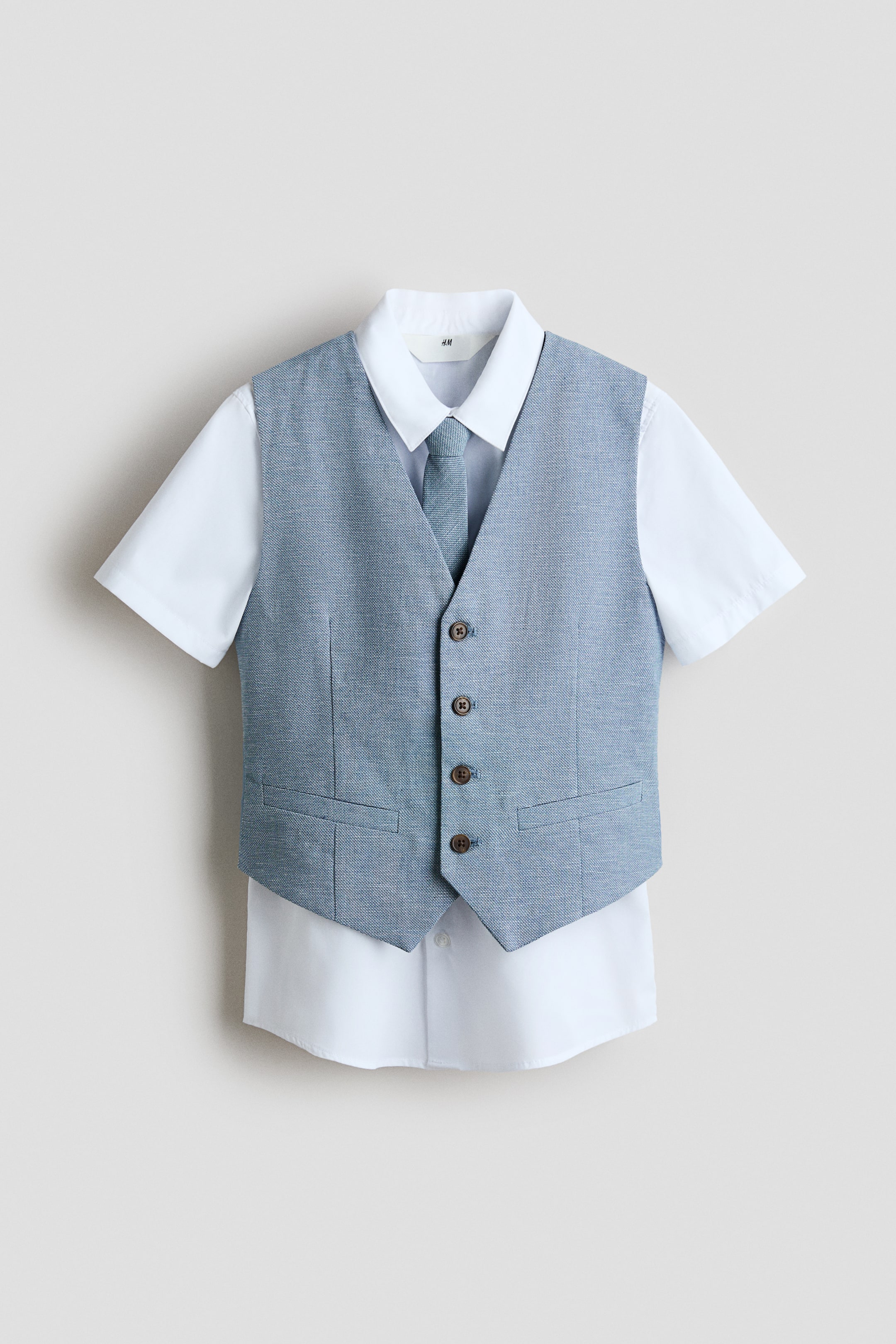 Agrandir l'image: Un ensemble enfant composé d’un gilet texturé bleu‑gris, d’une chemise blanche à manches courtes et d’une cravate assortie est présenté de face. Le gilet présente une encolure en V, quatre boutons marron sur le devant et deux fausses poches, superposé à une chemise blanche à col et manches courtes avec une cravate bleu‑gris assortie au col.