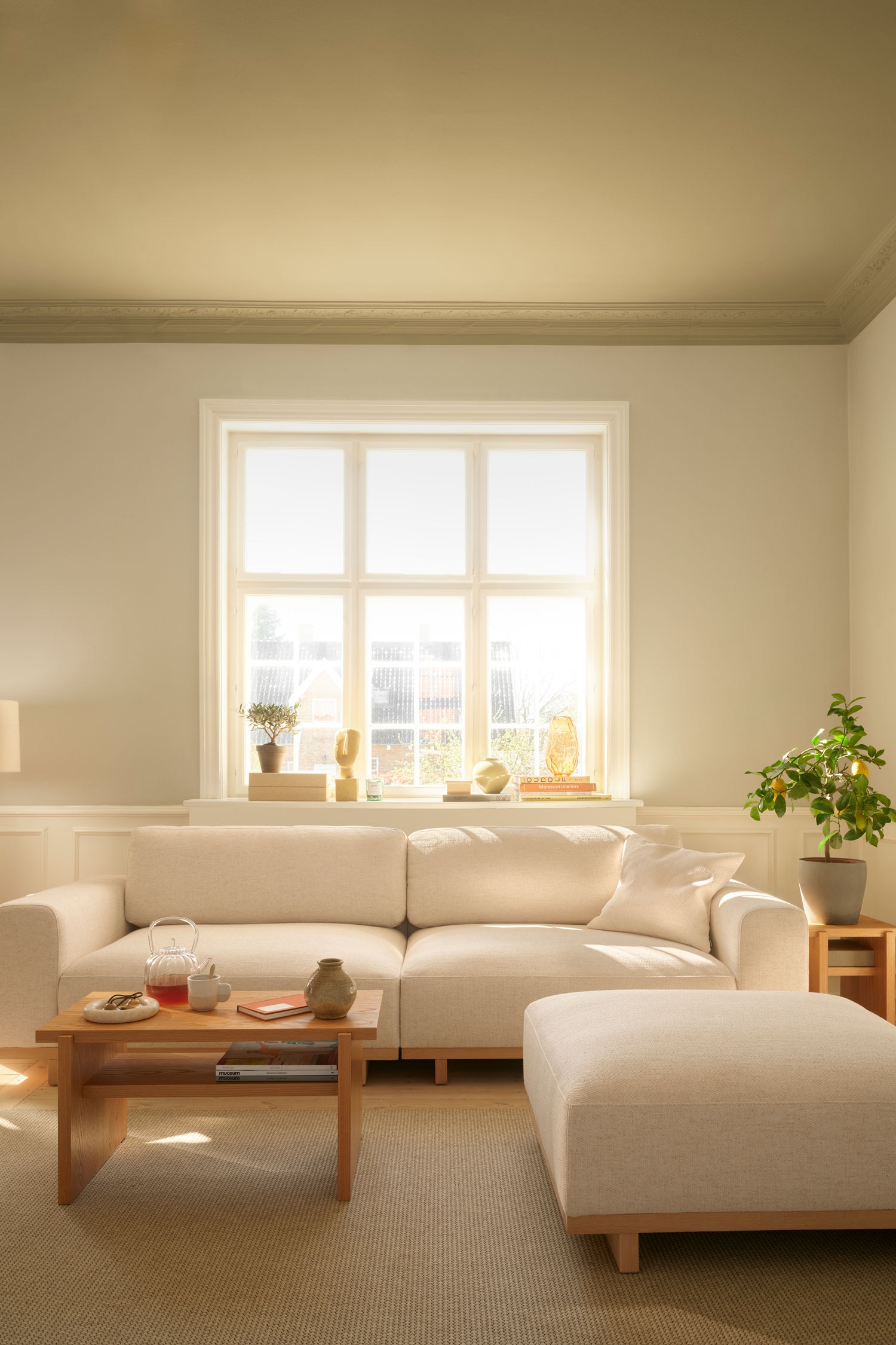Een lichtbeige gestoffeerde sofa staat naar voren gericht en heeft een laag profielontwerp met twee brede zitkussens, bijpassende rugkussens en dikke, blokvormige armleuningen. De sofa rust op een licht houten platformonderstel dat wordt ondersteund door korte, blokvormige poten.