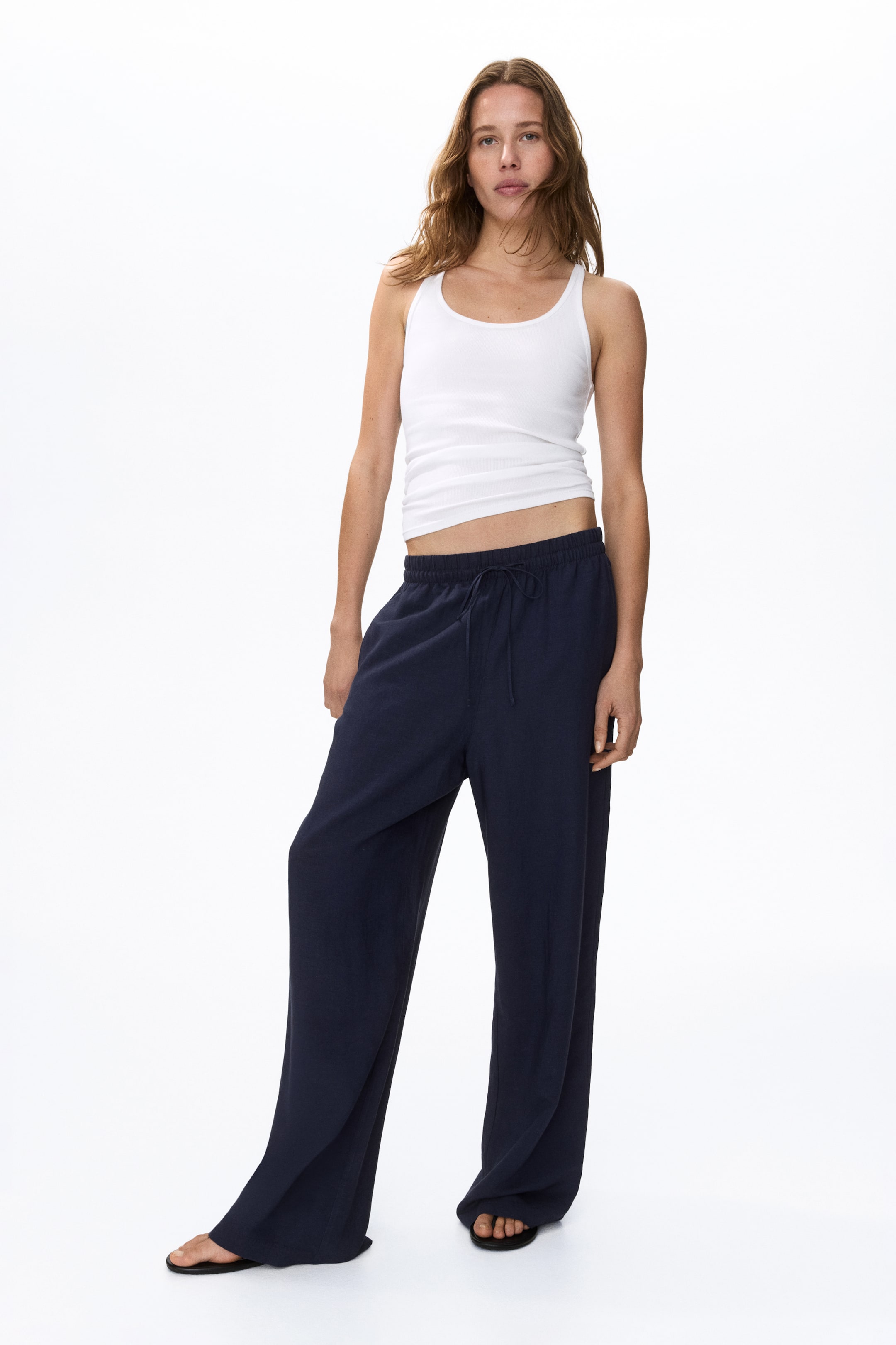 Agrandir l'image: Pantalon large bleu marine avec ceinture élastiquée à cordon de serrage et coupe ample et fluide, visible de face. Les jambes larges tombent librement, s'évasant légèrement sur des sandales plates noires, et sont présentées avec un débardeur côtelé blanc.