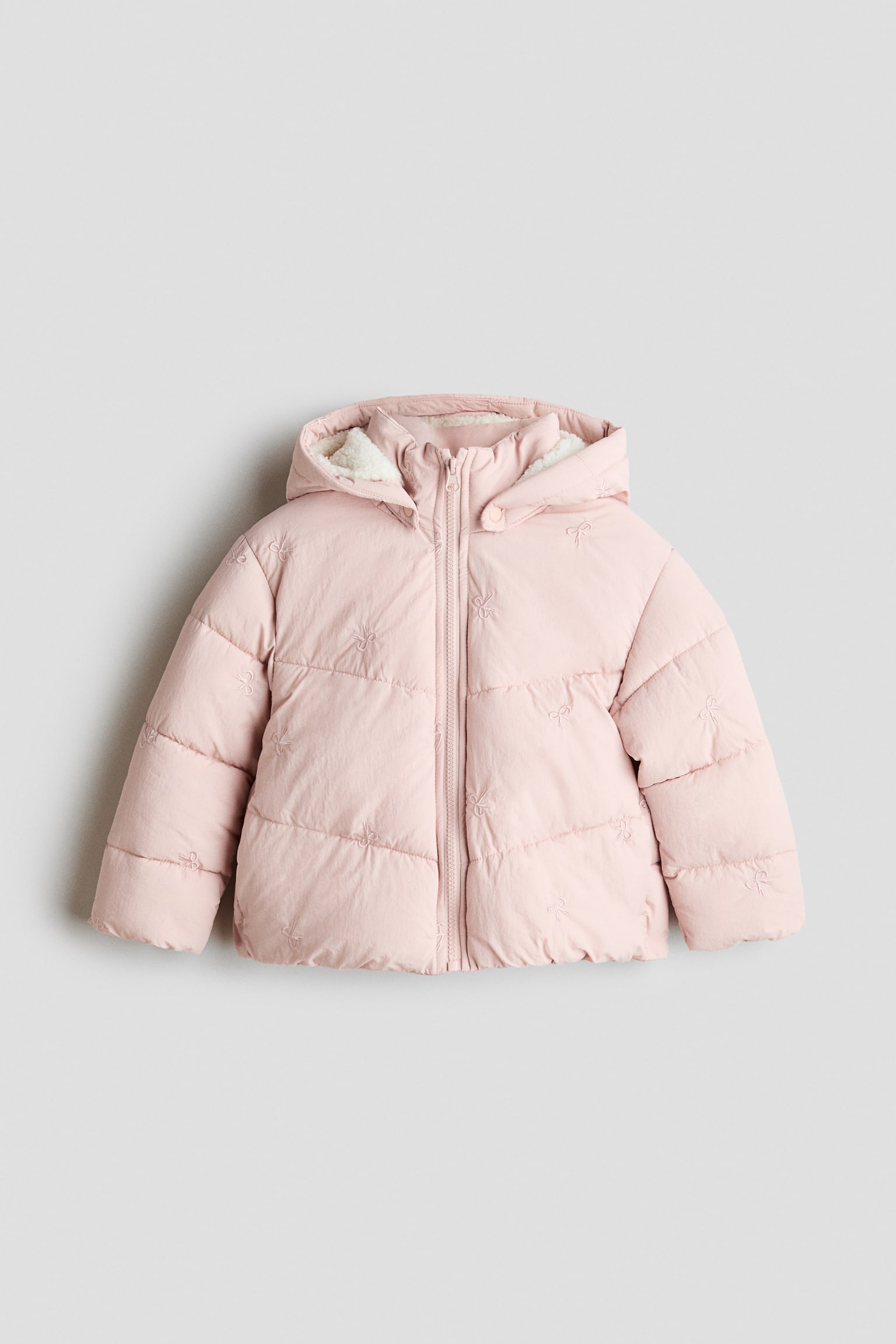 Una giacca imbottita rosa chiaro Ã¨ mostrata frontalmente, con chiusura frontale full zip e cappuccio integrato con fodera in pile bianco. La giacca presenta una trapuntatura orizzontale impreziosita da piccoli motivi a fiocco ricamati su tutta la superficie.