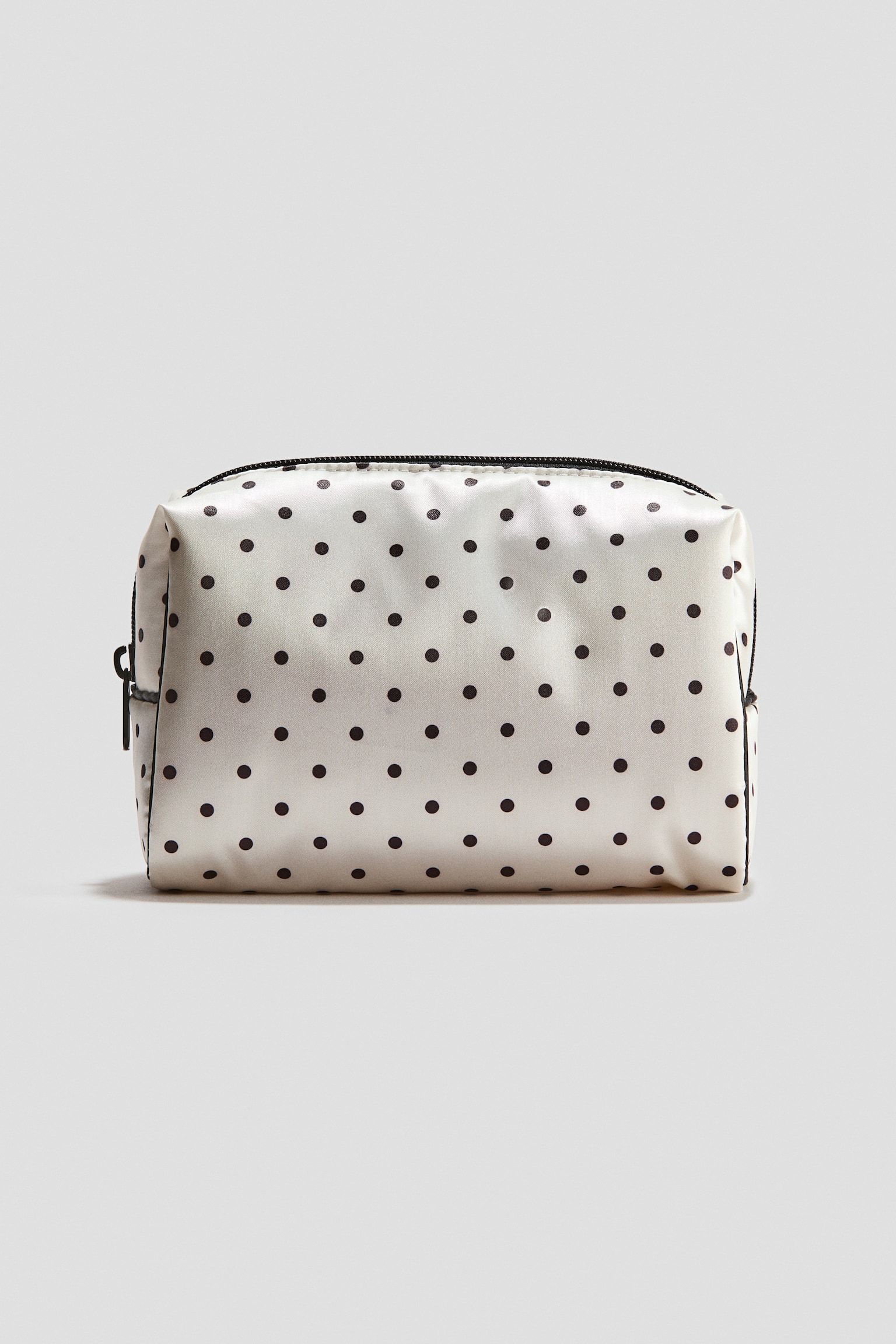A rectangular, soft-sided cosmetic bag made of off-white fabric with a black polka dot pattern is standing upright in a front-facing view. The bag is featuring a black zipper closure along the top with a black zipper pull on the side.