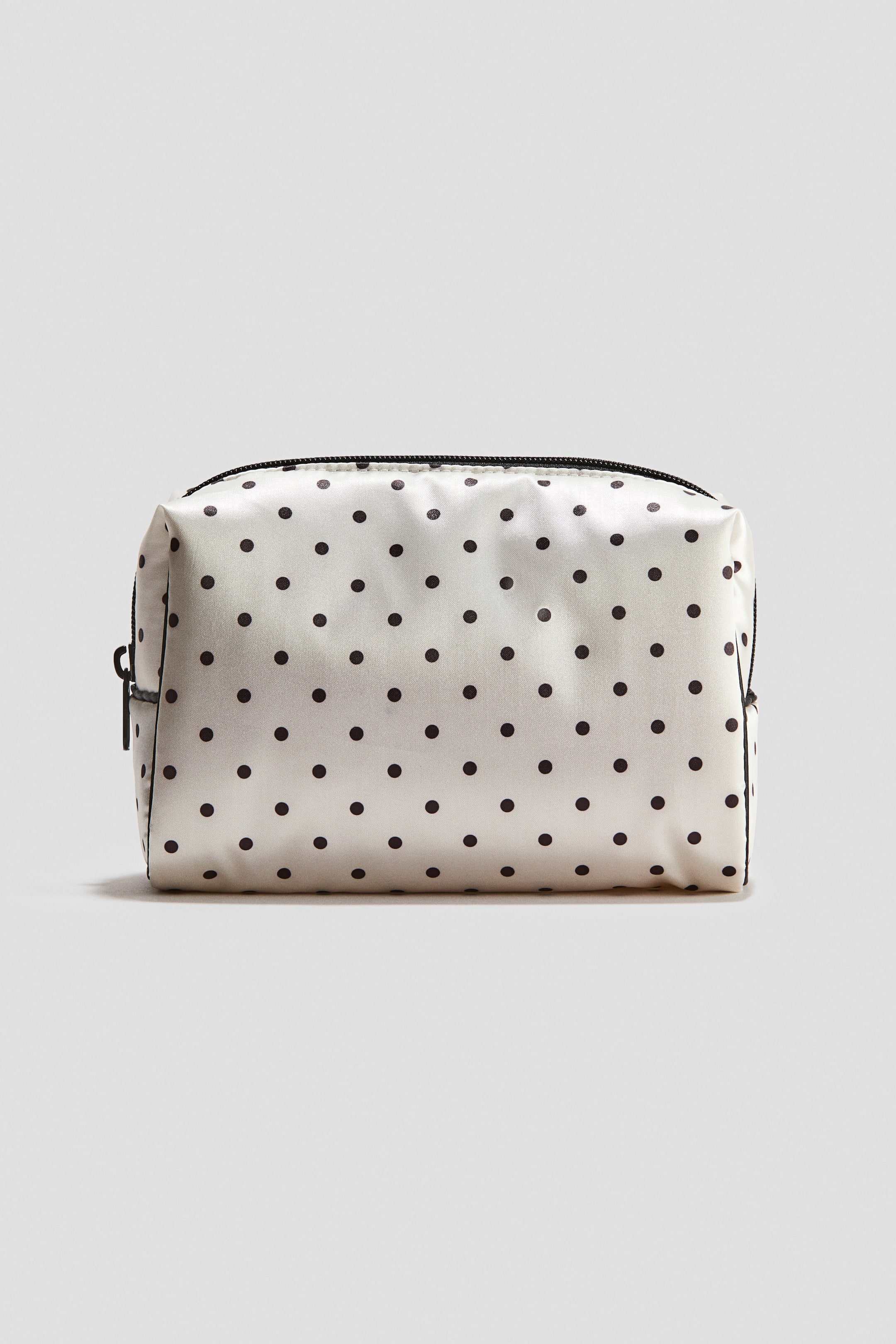 View larger image: A rectangular, soft-sided cosmetic bag made of off-white fabric with a black polka dot pattern is standing upright in a front-facing view. The bag features a black zipper closure along the top with a black zipper pull on the side.