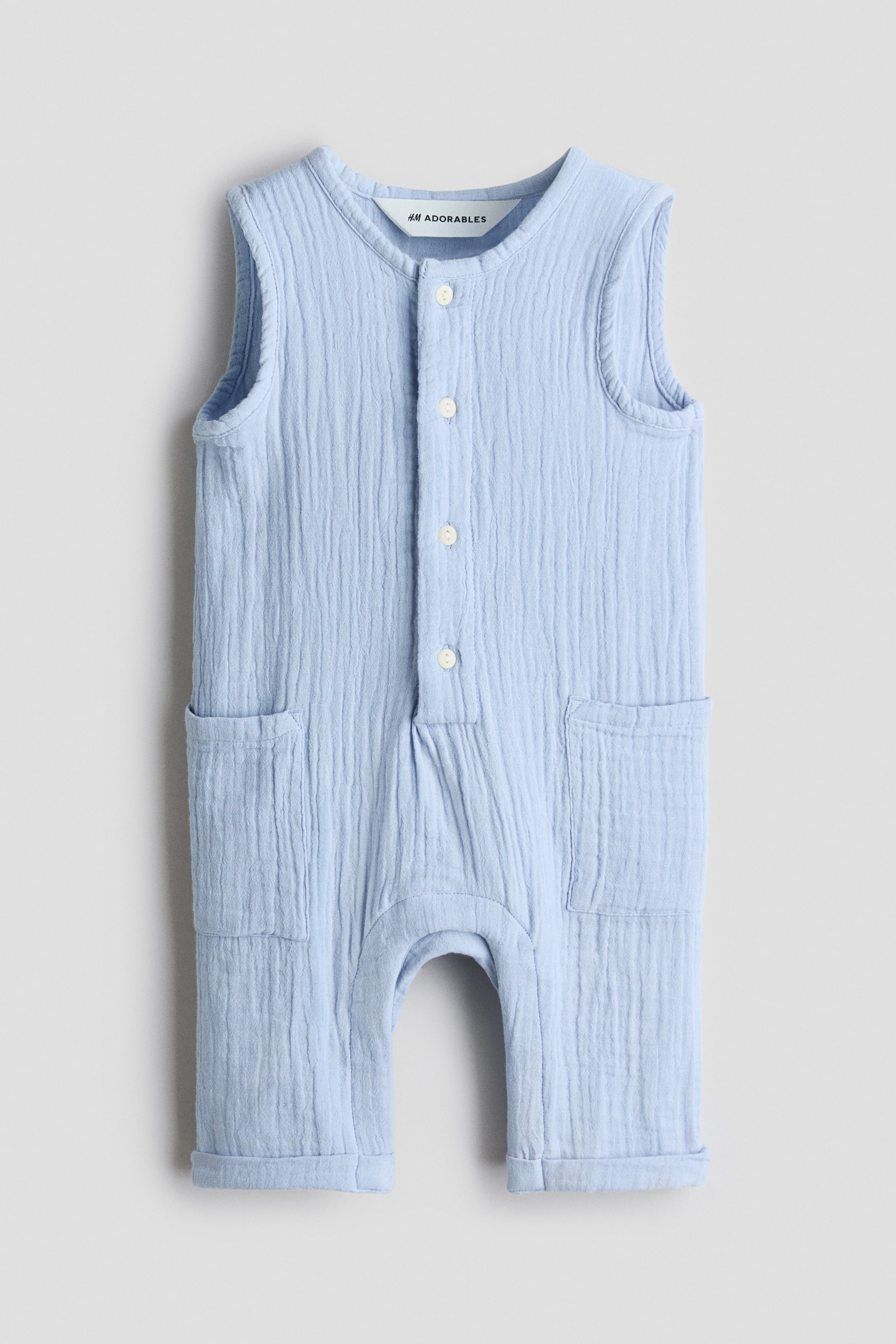 Agrandir l'image: Une combinaison sans manches en mousseline froissée bleu clair de la collection Adorables d'H&M est présentée de face, avec un col rond, des boutons sur le devant et deux poches plaquées ouvertes sur les côtés. Les ourlets des jambes montrent des revers cousus.