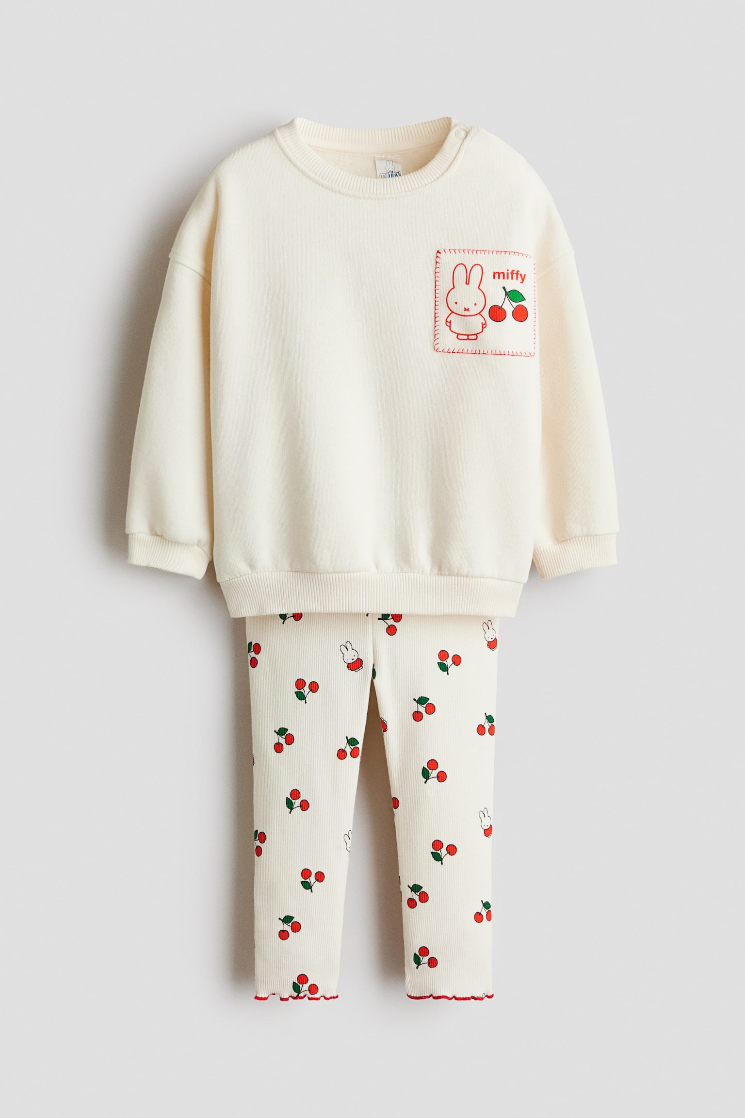 A front-facing, two-piece childrenâs set is showcasing an off-white sweatshirt featuring a ribbed crew neck, ribbed cuffs, a ribbed hem, and a chest patch pocket with a Miffy and cherry graphic. The matching ribbed leggings are displaying an all-over print of red cherries and small Miffy illustrations, finished with red-trimmed lettuce-edge hems.