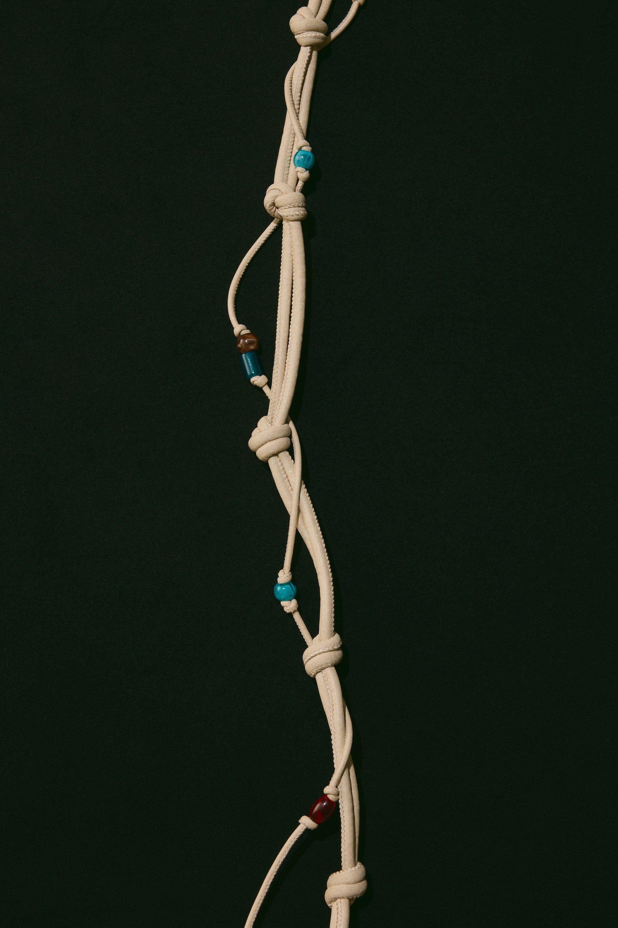 View larger image: A flat beige cord hangs vertically, showcasing several knots along its length. Adorning the cord are various beads, including round turquoise, brown barrel, blue cylindrical, and red oval beads. These are attached to thinner cords that intertwine with the main cord.