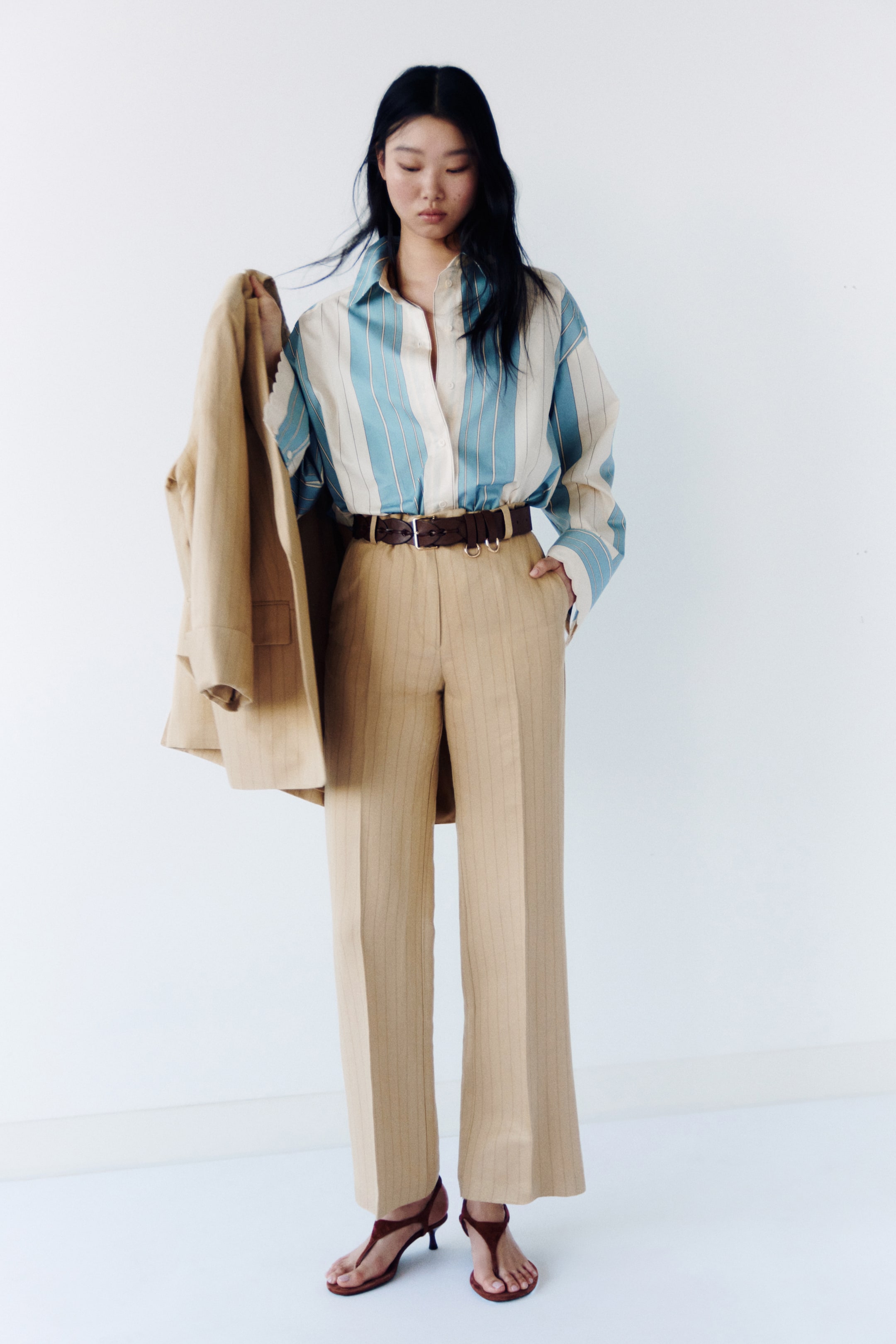 Ver imagen más grande: El producto es una camisa de botones con rayas en crema y azul claro, con un cuello en punta, metida en unos pantalones de talle alto color beige con pinzas y pernera ancha, ajustados con un cinturón de anilla en D de color marrón oscuro. Se lleva una americana a juego con rayas beige sobre un brazo, y unas sandalias de tiras en T de color rojo oscuro con tacón bajo.