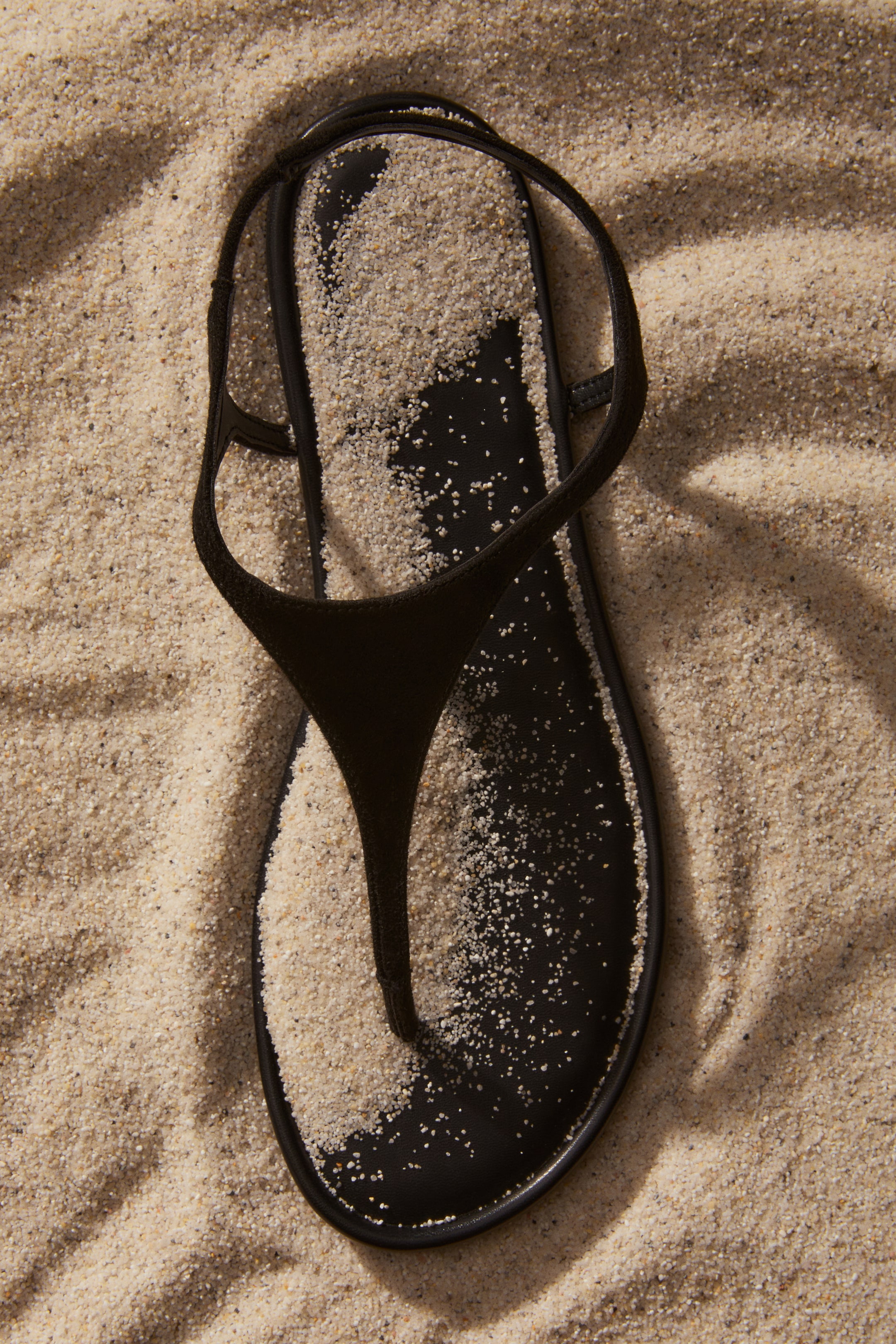 View larger image: A black thong sandal is lying with its toe pointing upwards, featuring thin straps and a black sole partially covered in fine sand.