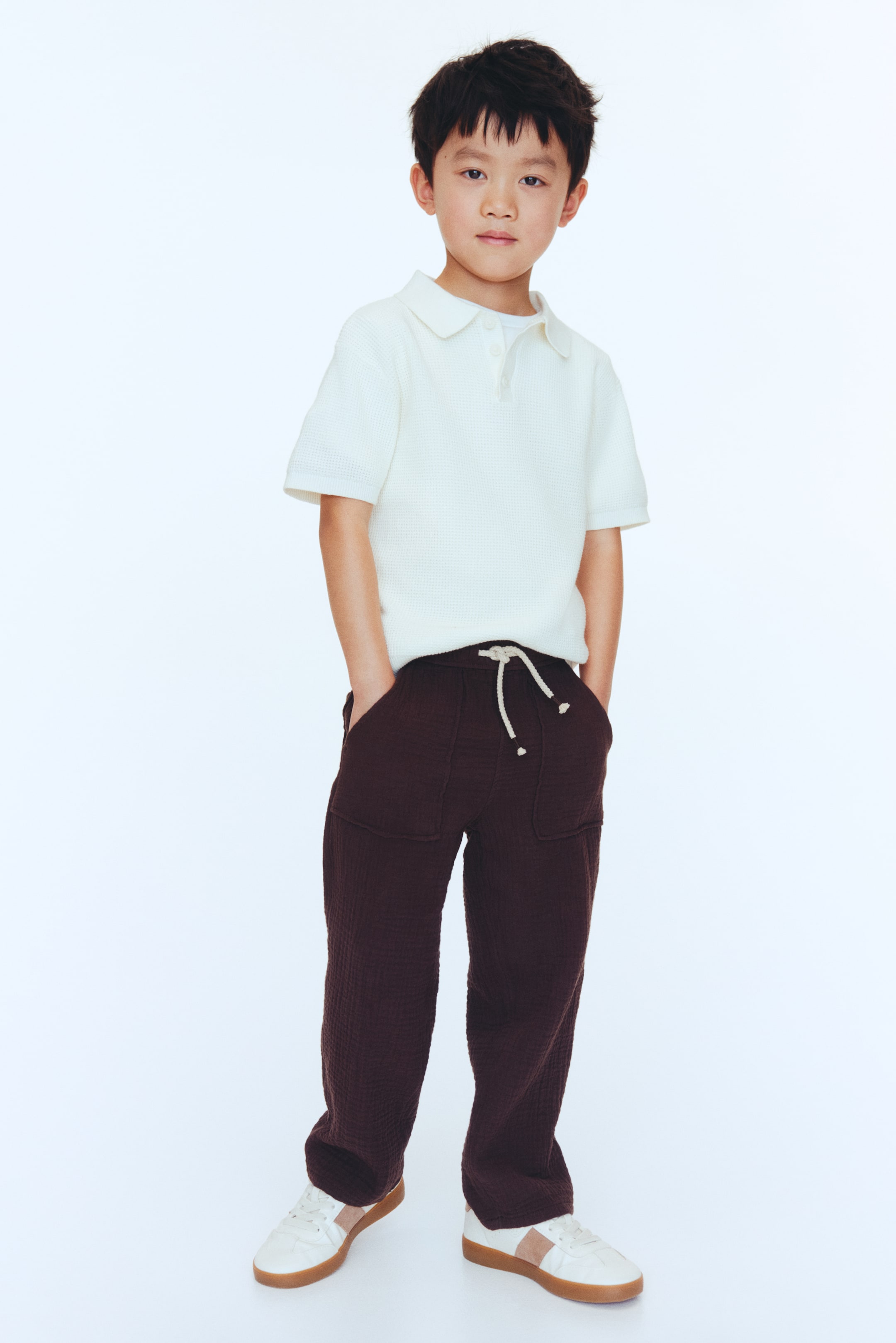 View larger image: An off-white textured short-sleeve polo shirt with a collar and two-button placket is being worn. Dark brown textured pants, featuring a drawstring waist with a light-colored tie and front patch pockets, are paired with white sneakers showcasing light brown side stripes and gum soles.